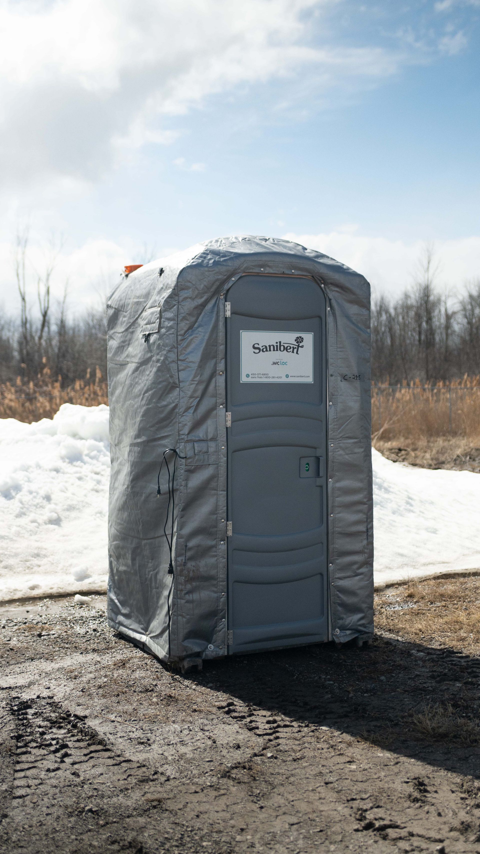 Toilettes portables grises sur sol en terre battue à l'extérieur.