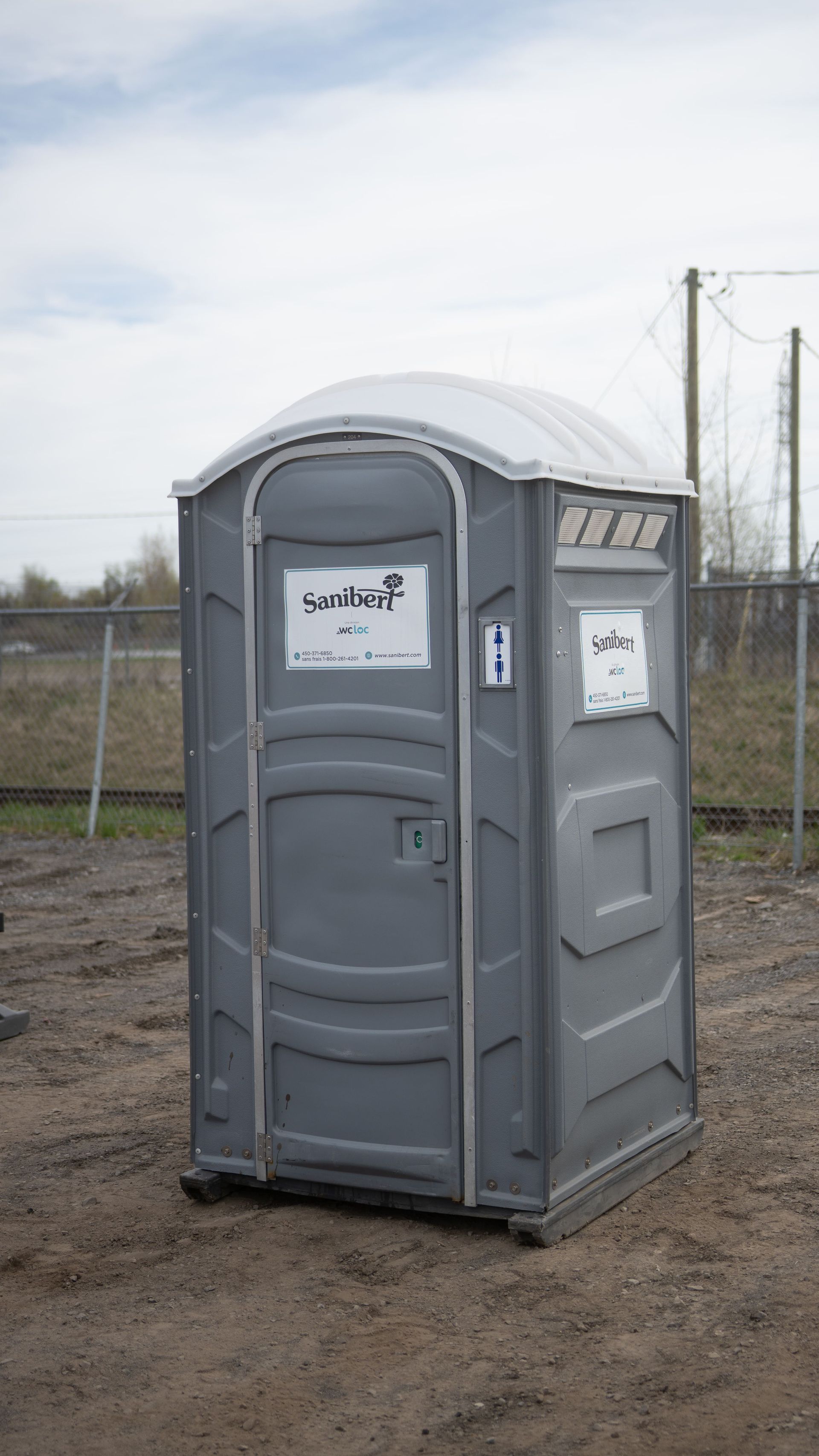 Toilettes portables grises sur sol en terre battue à l'extérieur.