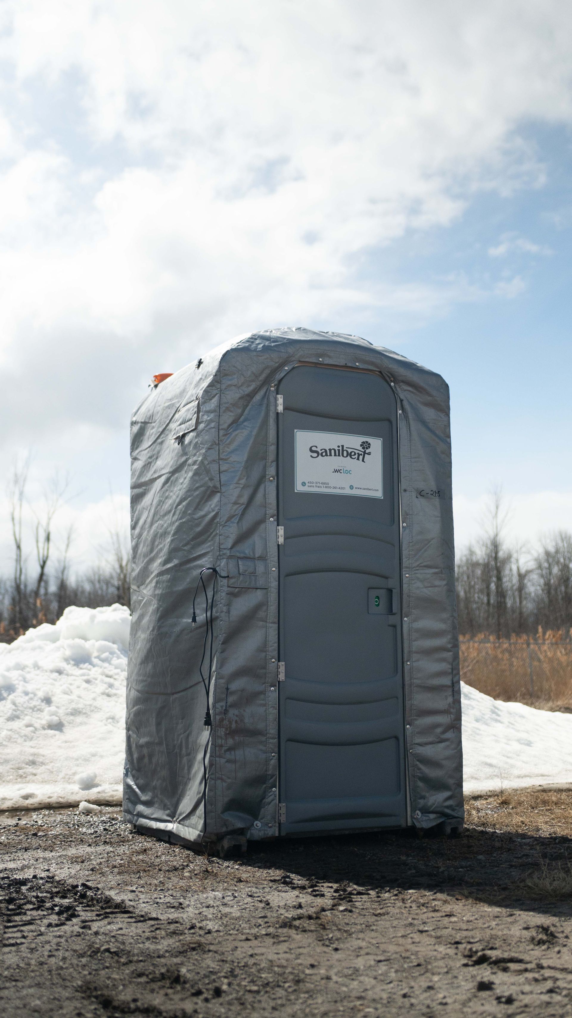 Toilettes portables grises sur un chemin de terre, avec neige et arbres en arrière-plan. Ciel nuageux.