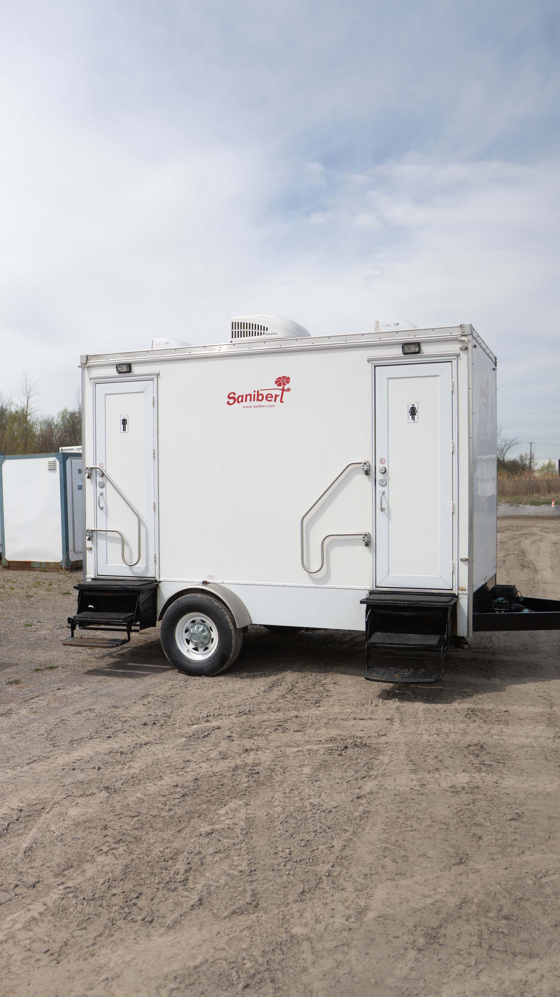 Remorque de toilettes portables blanches avec deux entrées séparées sur une surface en terre.