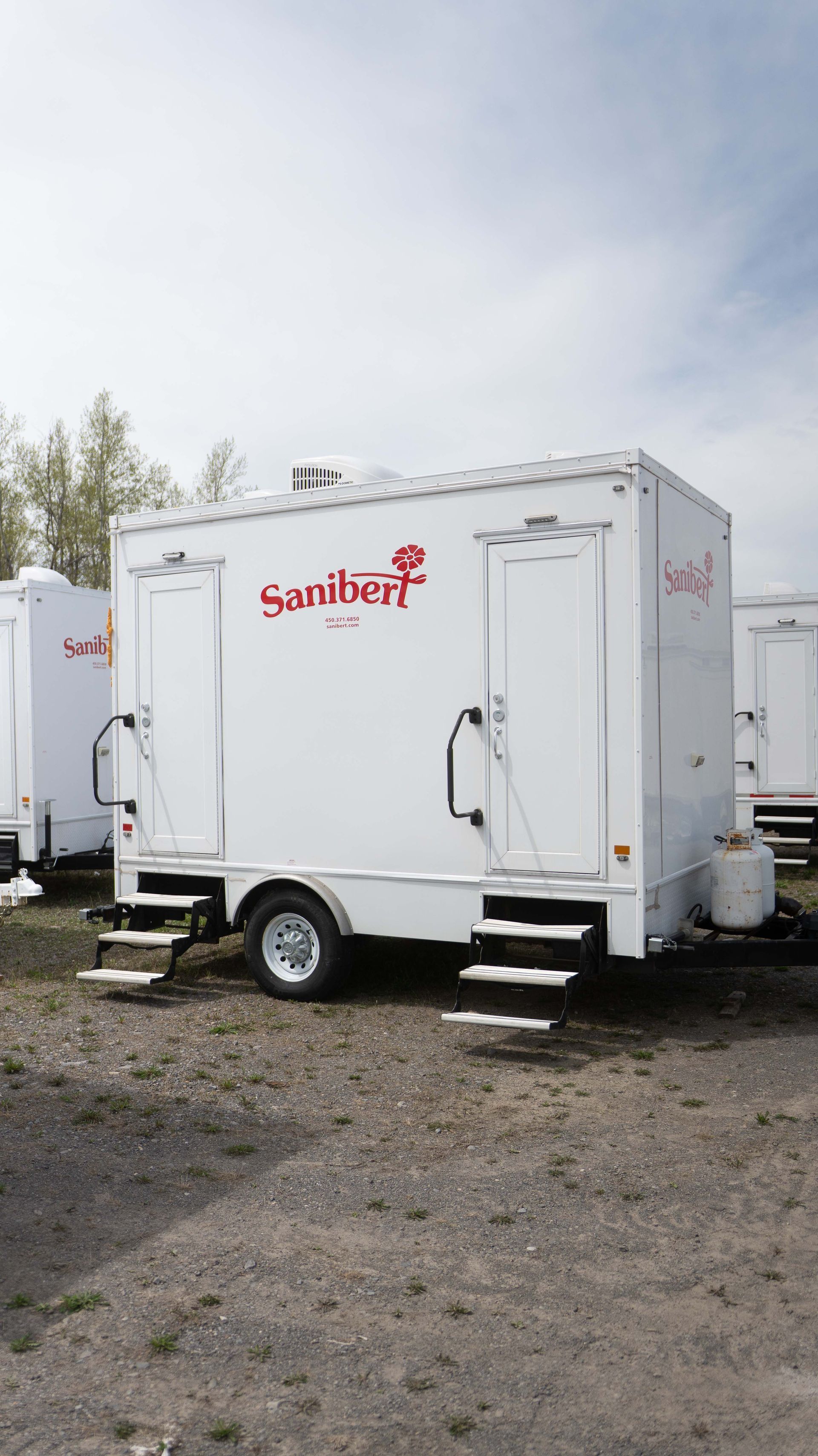 Remorque de toilettes portables blanches avec deux portes et marches, stationnée à l'extérieur.