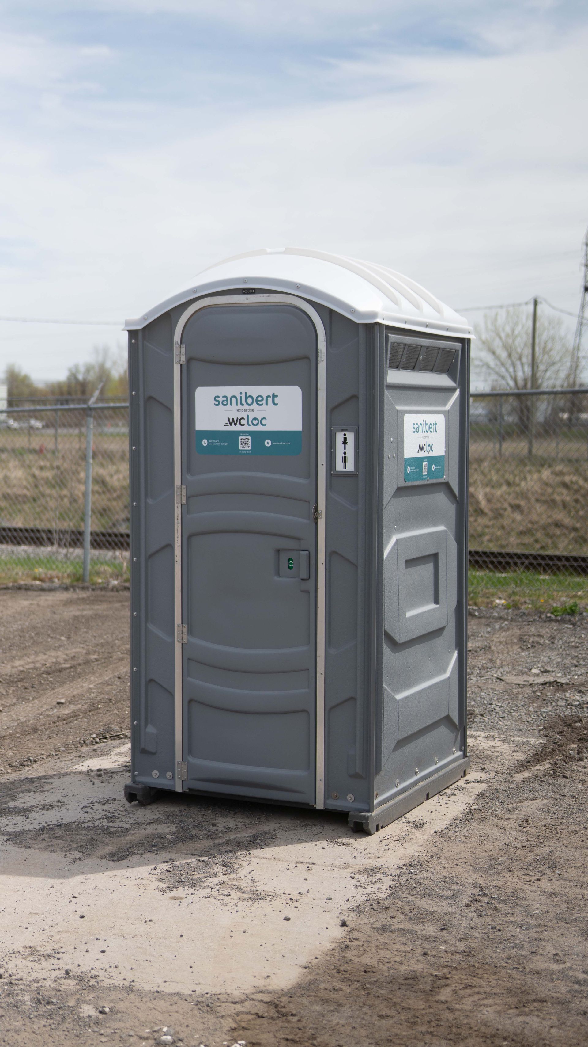 Toilettes portables grises dans un terrain en gravier avec une clôture grillagée et un arrière-plan industriel.