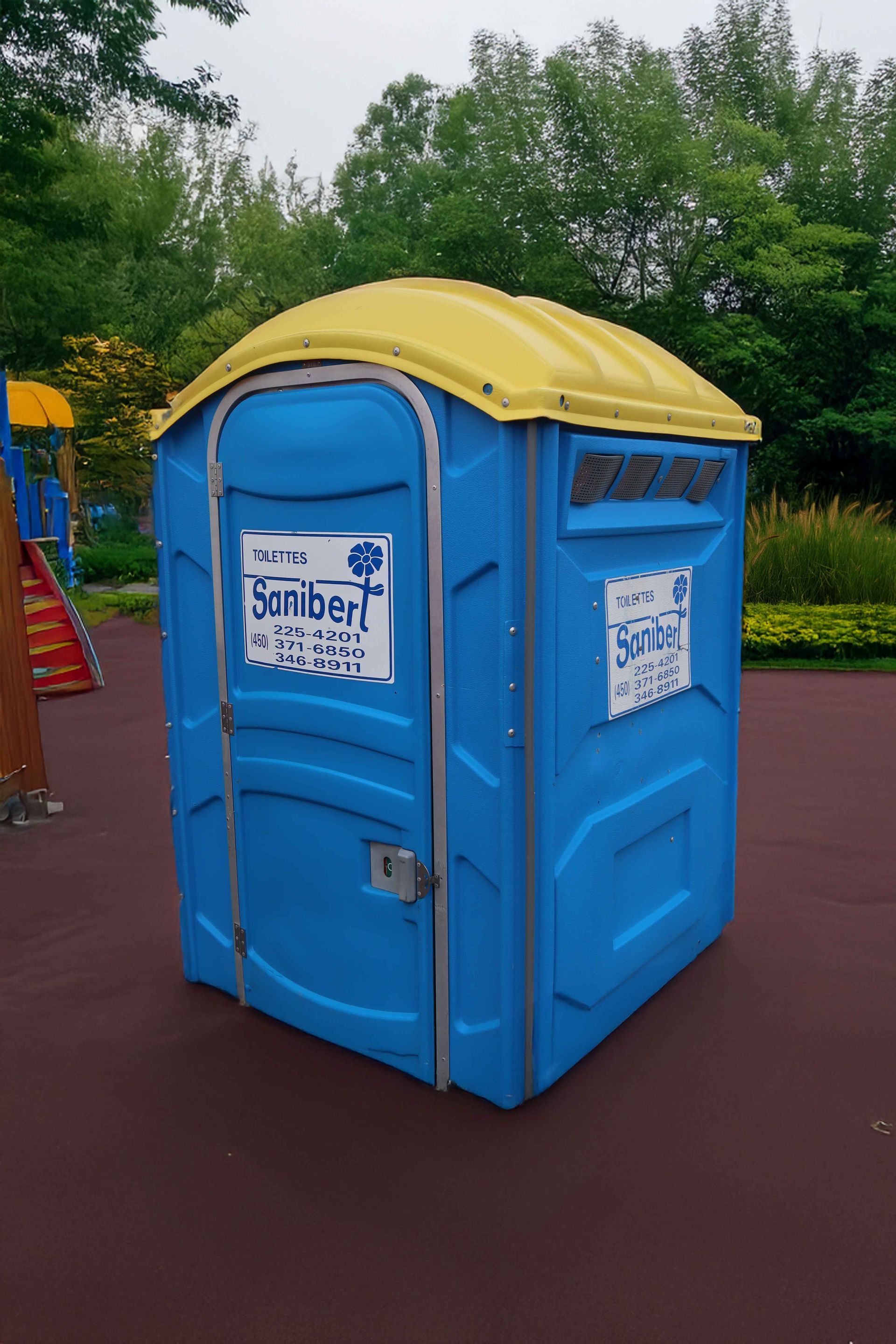 Toilettes portables bleues et jaunes sur une surface rouge dans un parc, avec des équipements de jeux en arrière-plan.