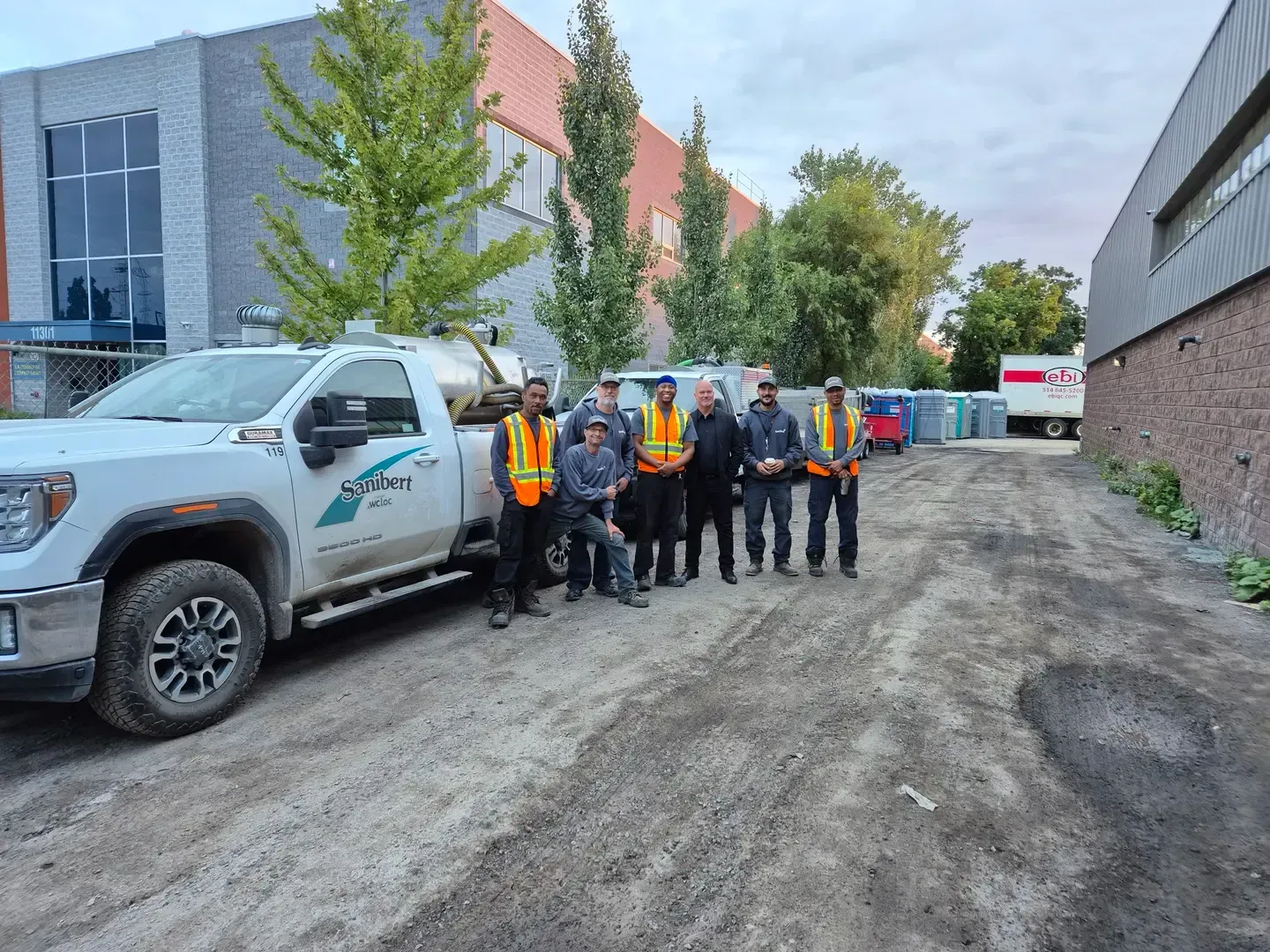 Un groupe d'hommes portant des gilets de sécurité pose à côté d'une camionnette blanche, devant un bâtiment.