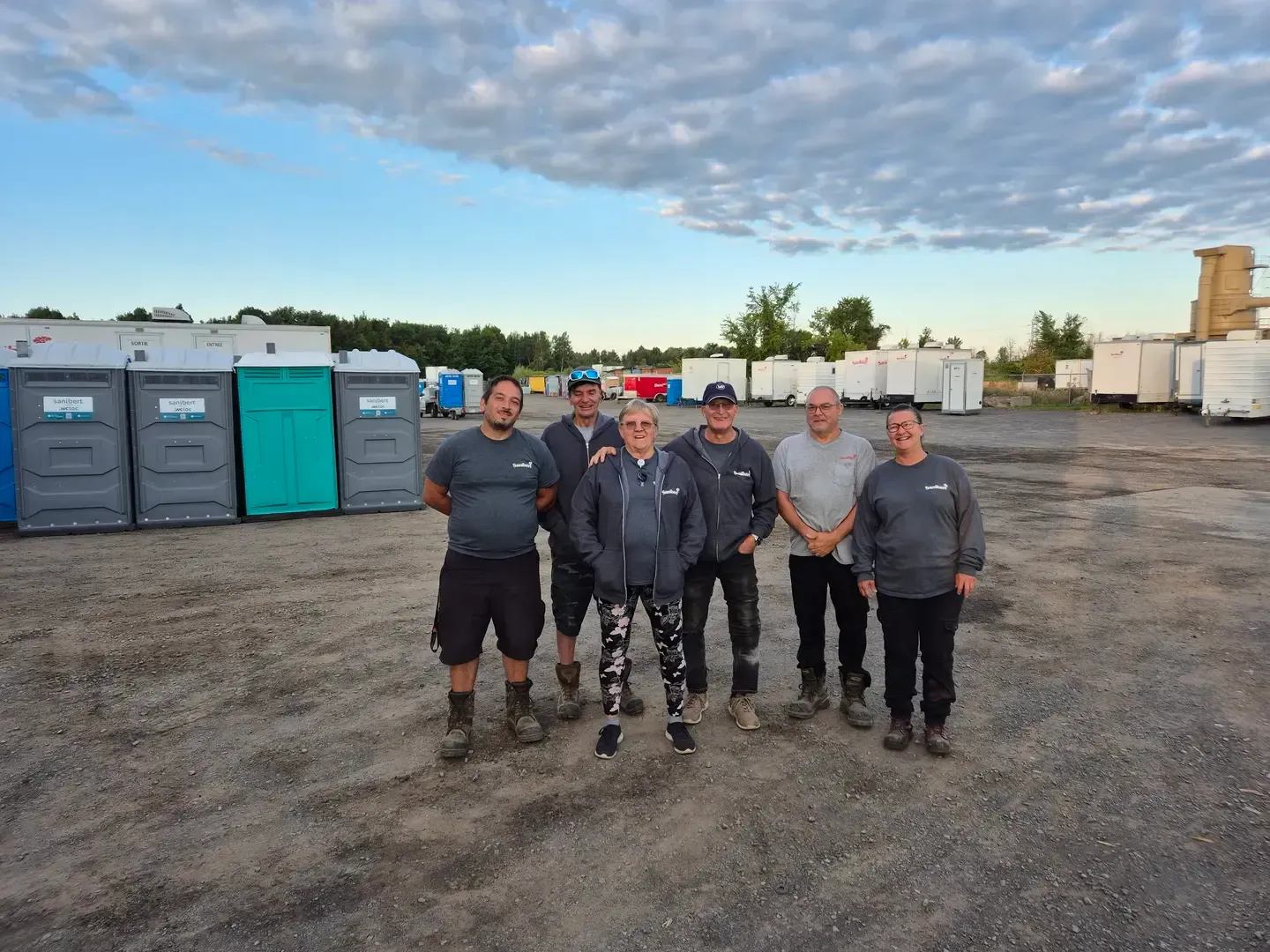 Groupe de sept personnes posant à l'extérieur, debout devant des toilettes portables dans un terrain en gravier.