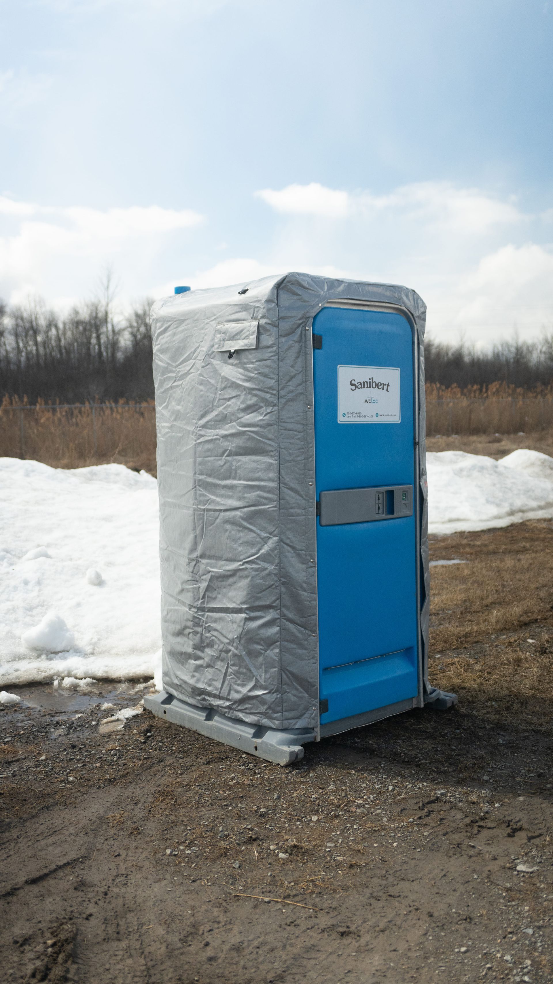 Des toilettes portables bleues, recouvertes d'une bâche grise, sont posées sur un sol gravillonné à côté d'un sol enneigé.