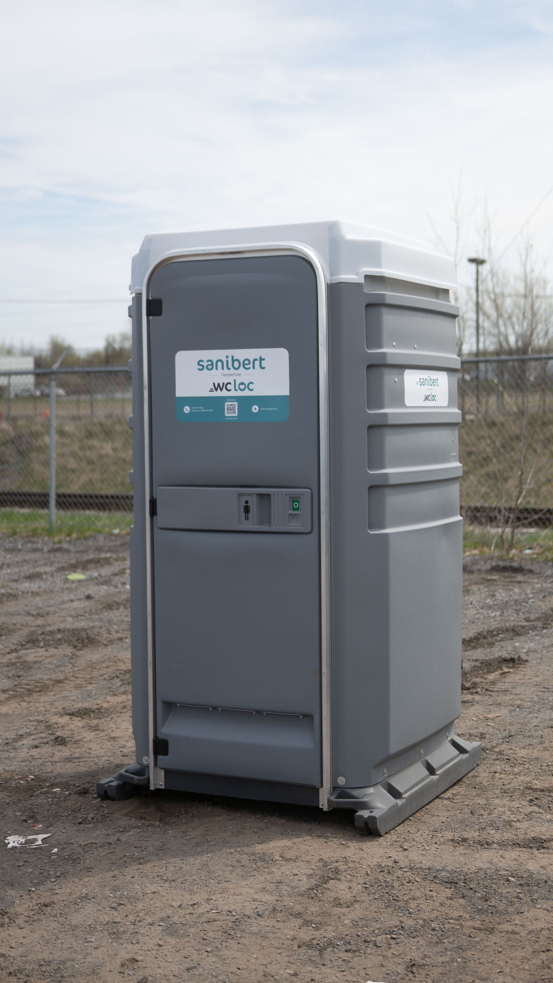 Des toilettes portables grises avec un panneau se trouvent à l'extérieur sur une surface en terre.