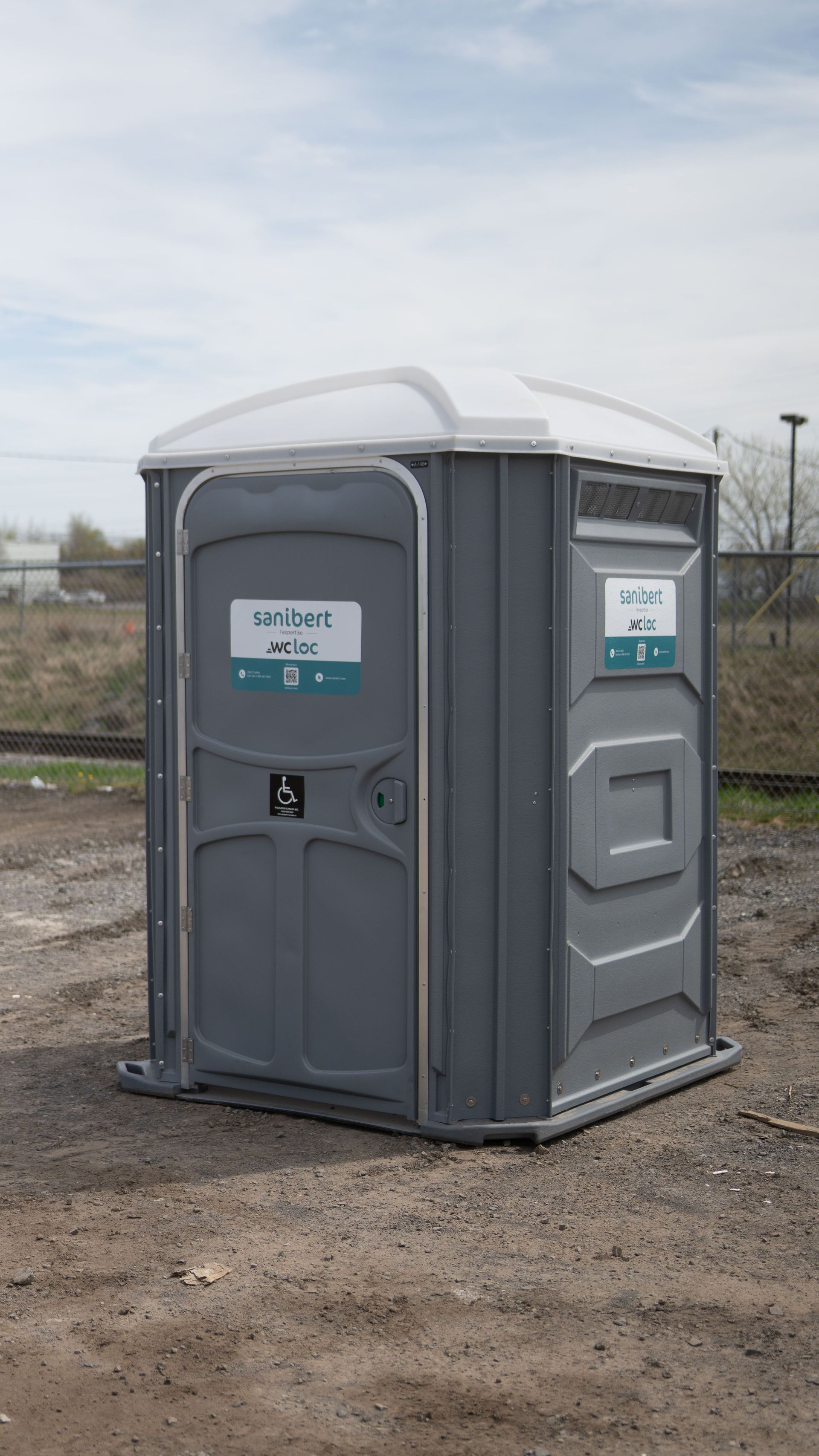 Toilettes portables grises sur gravier, en extérieur. Un panneau indique leur accès. Ciel nuageux en arrière-plan.