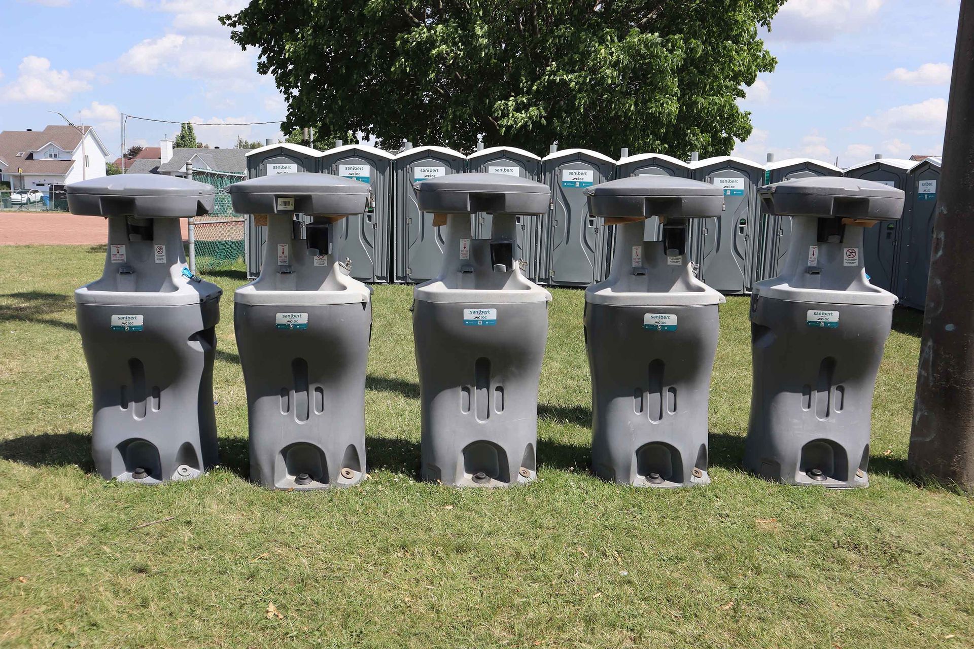 Cinq stations de lavage des mains portables grises devant une rangée de toilettes portables sur l'herbe, journée ensoleillée.