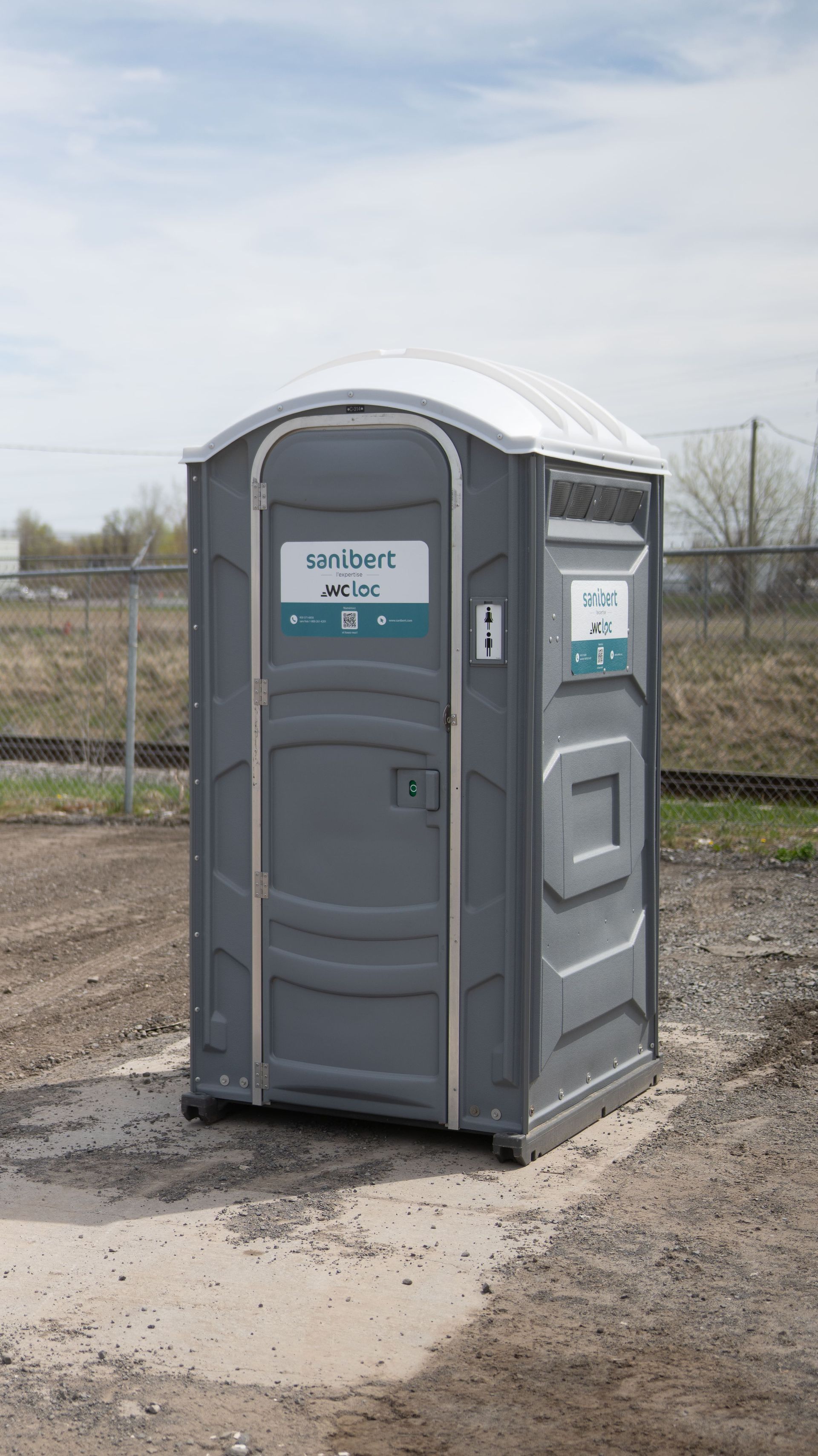 Toilettes portables grises sur gravier avec une clôture grillagée et des voies ferrées en arrière-plan.