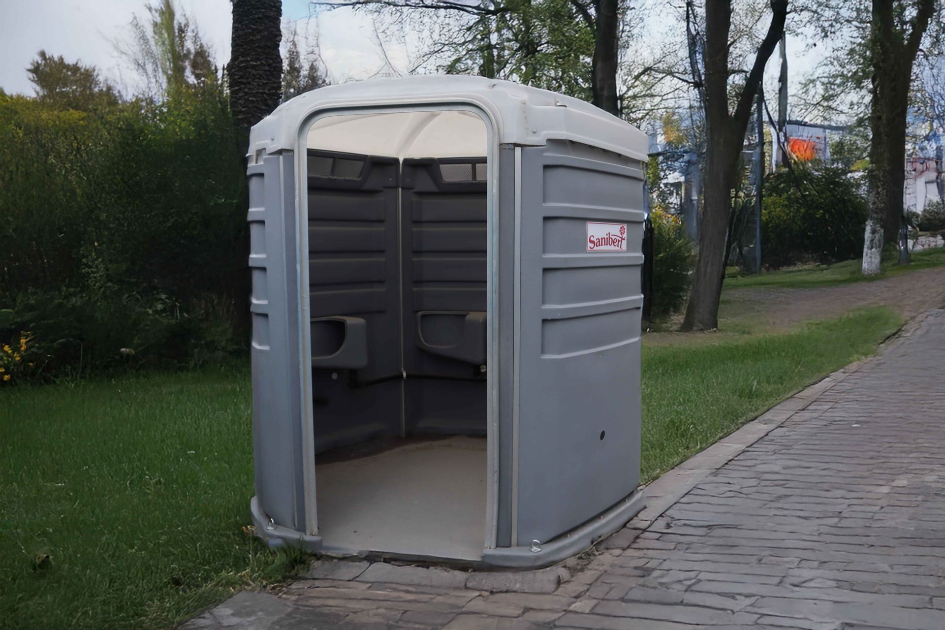 Toilettes portables grises sur un chemin pavé dans un parc avec de l'herbe et des arbres.