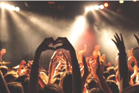Public au concert, bras levés, une personne formant un cœur avec ses mains. Lumières de la scène en arrière-plan.