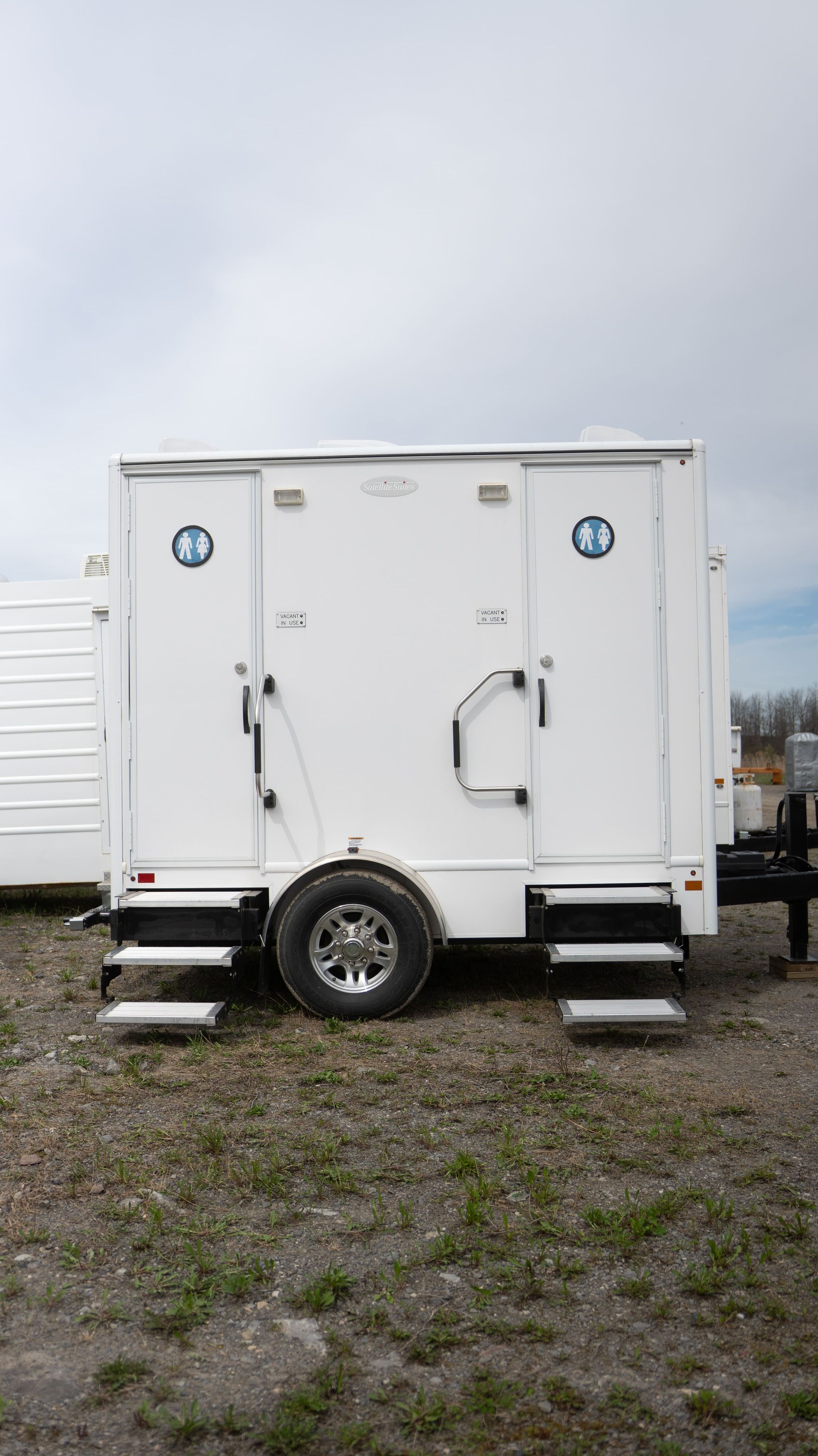 Remorque de toilettes portables blanches avec deux portes, marches et un pneu, à l'extérieur.
