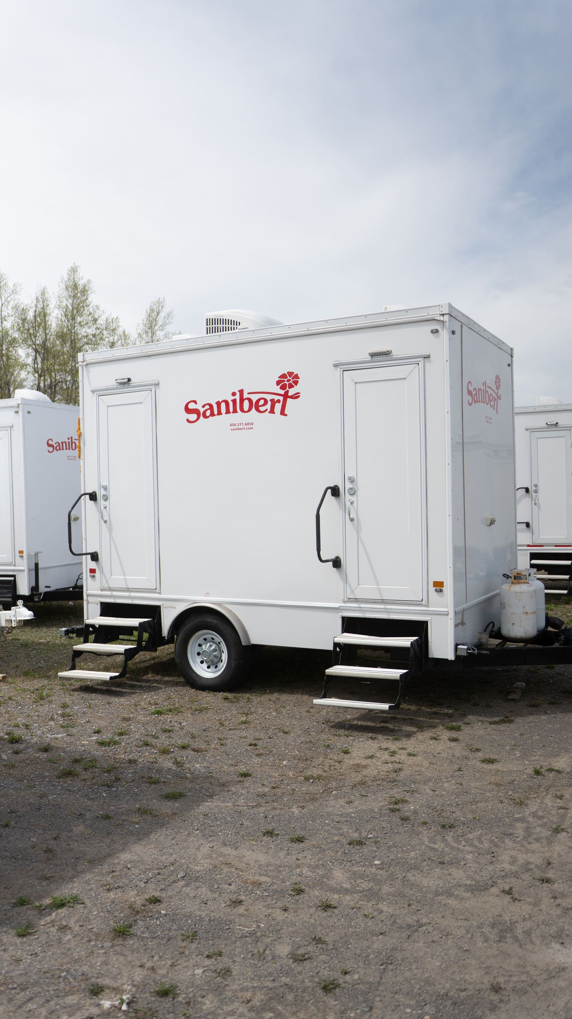 Remorque sanitaire portable blanche avec logo Sanibon, deux entrées, marches et une roue.