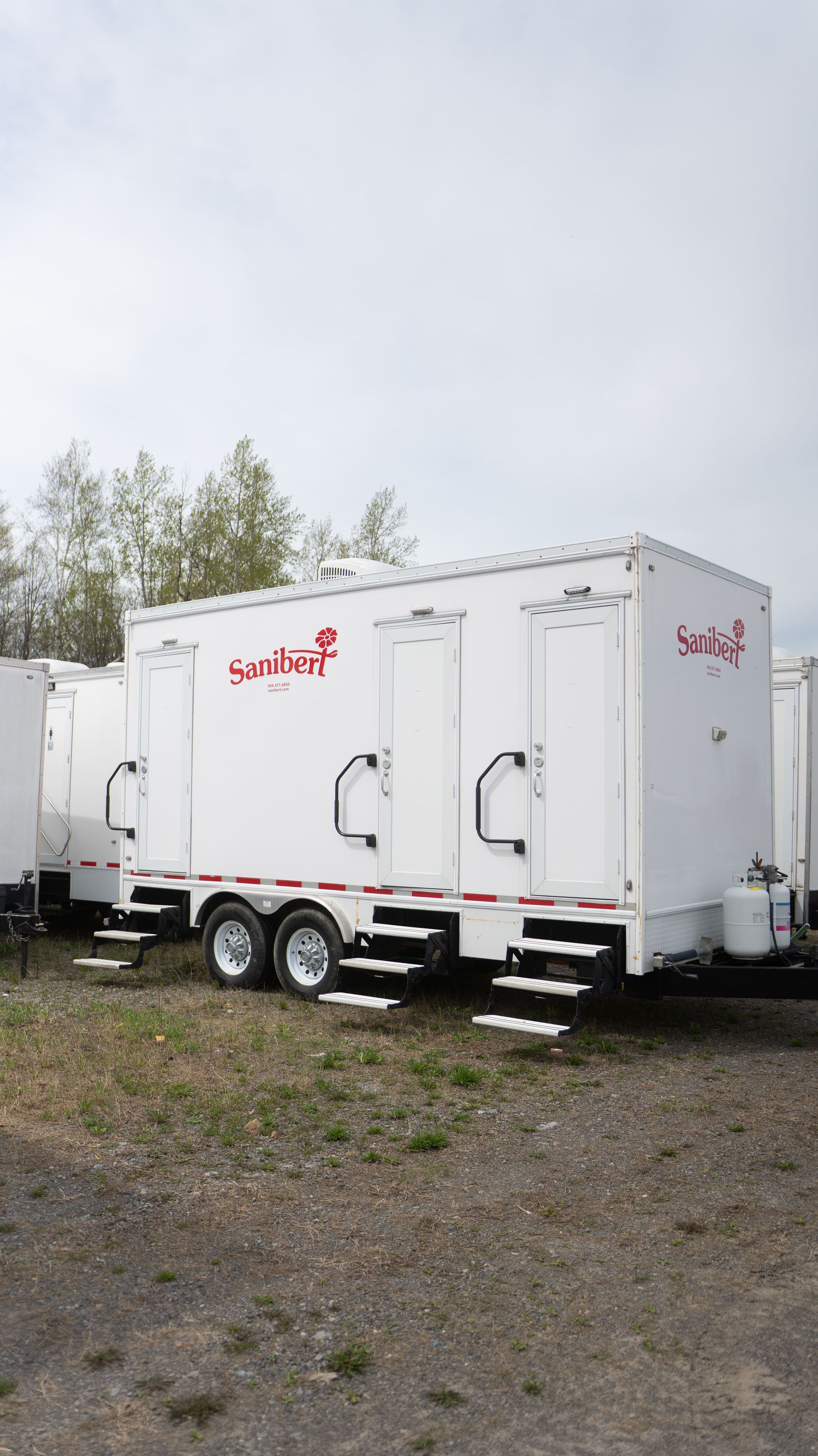 Toilettes mobiles blanches avec marches, garées en extérieur. Logo « Sanibell » en rouge.