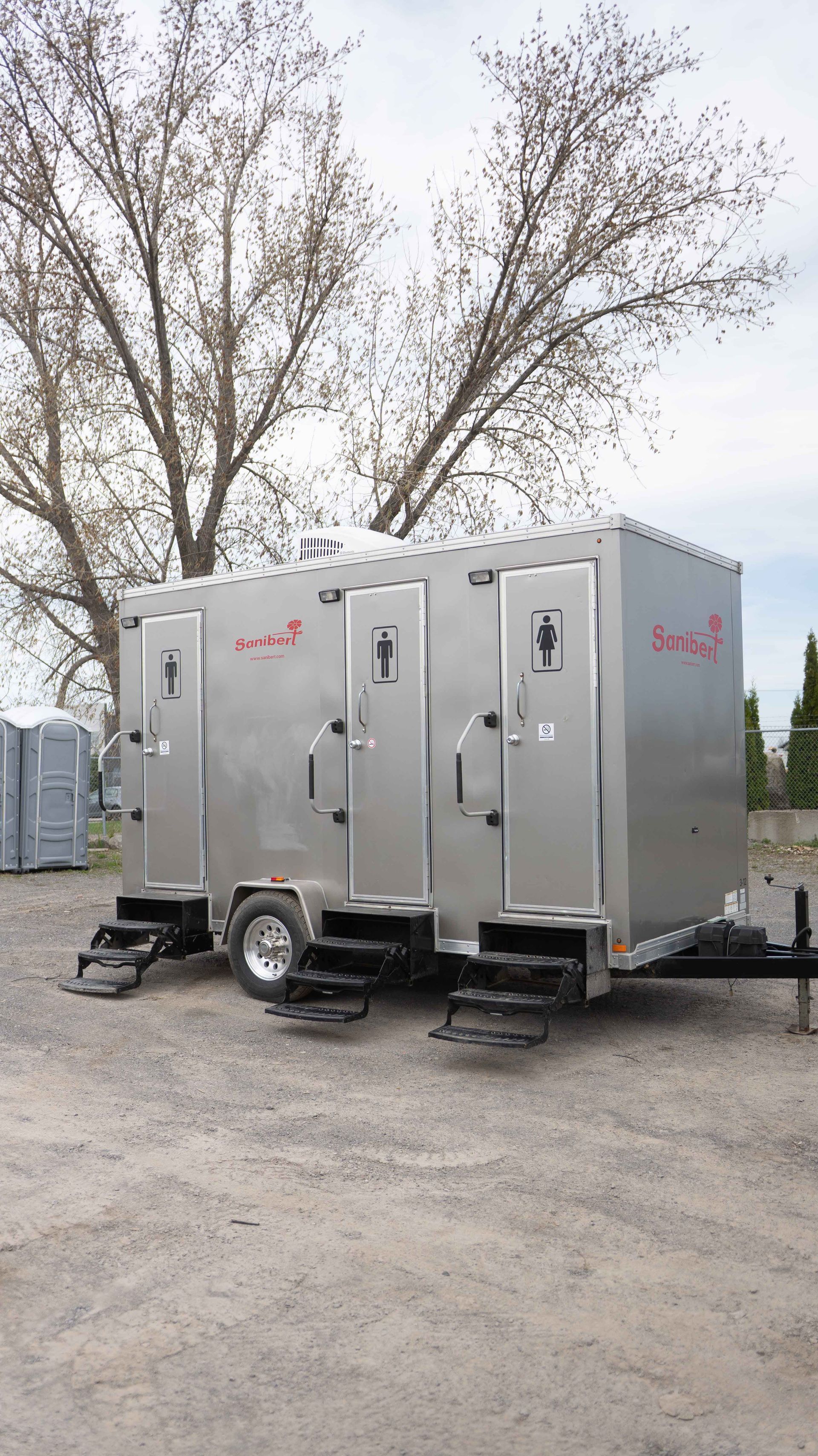 Remorque de toilettes portables argentées avec trois portes sur un terrain en gravier.