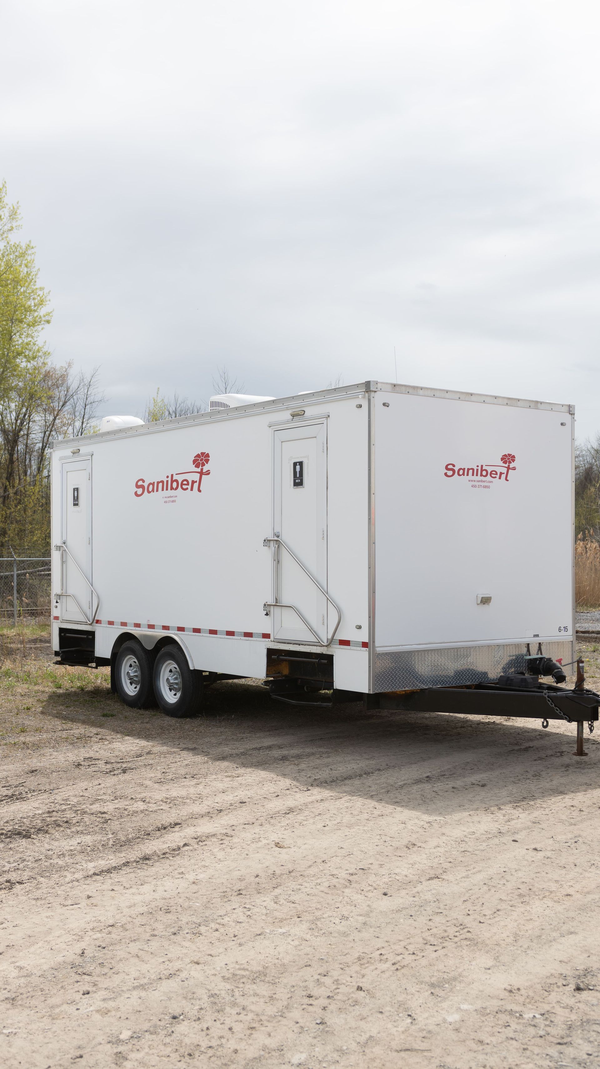 Remorque de toilettes portables blanches sur un terrain en gravier avec lettrage rouge.