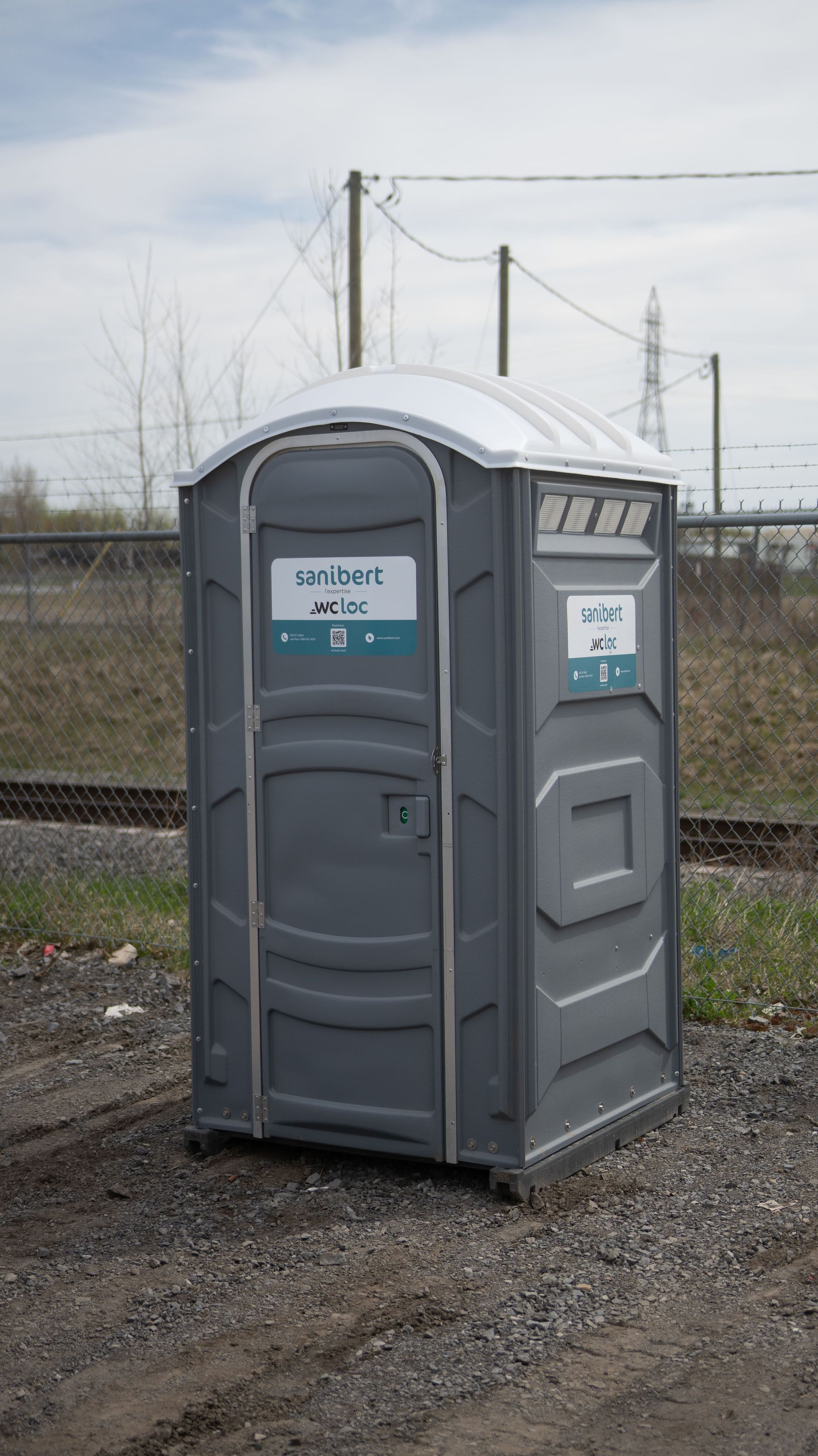 Toilettes portables grises sur gravier, près des voies ferrées.