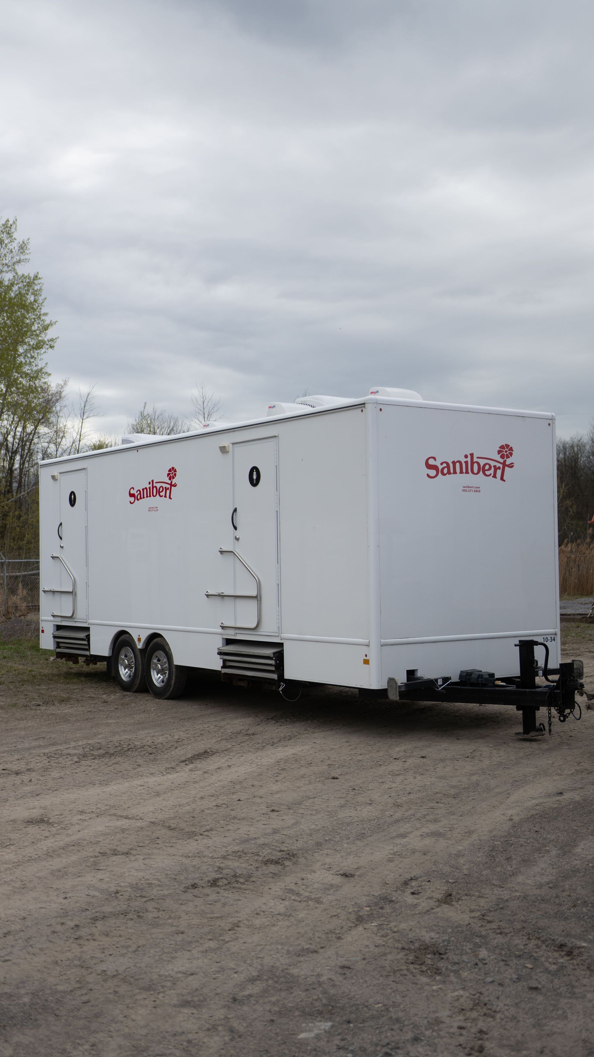Remorque de toilettes portables blanches avec logo rouge sur un terrain en gravier sous un ciel nuageux.
