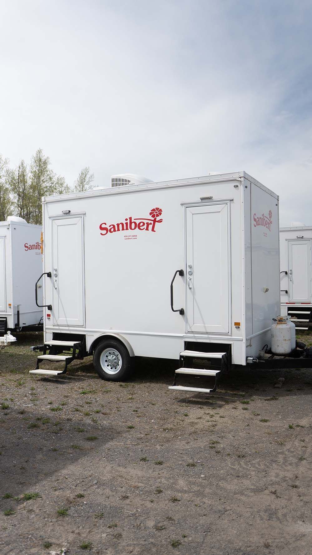 Toilettes mobiles blanches avec marches, sur gravier, sous un ciel nuageux. Logo rouge sur le côté.