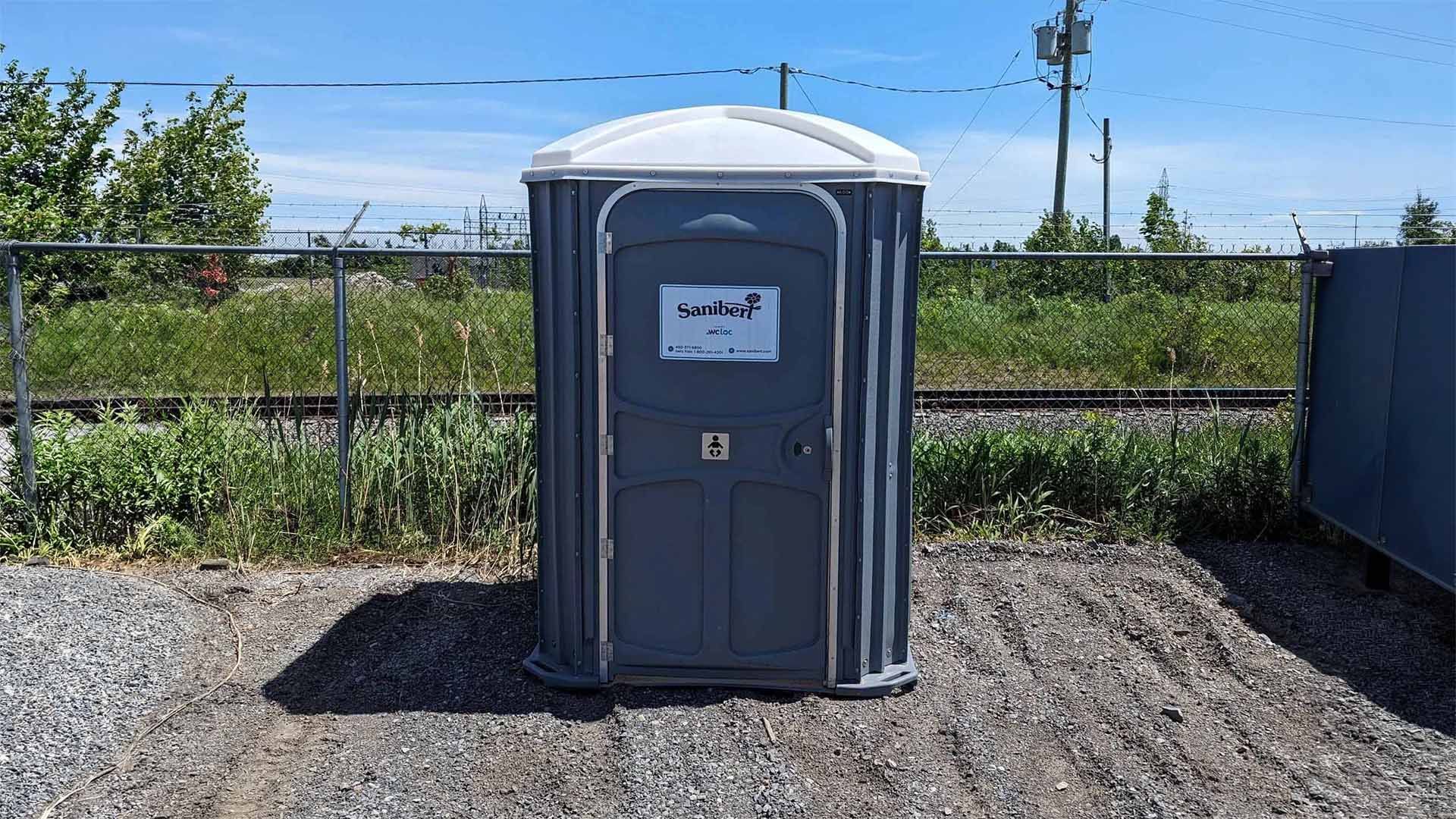 Toilettes portables grises sur gravier à côté d'une voie ferrée sous un ciel bleu.