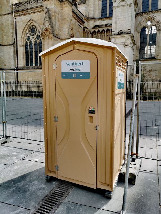 Toilettes portables beiges devant un bâtiment en pierre avec des fenêtres cintrées, sur une zone pavée.