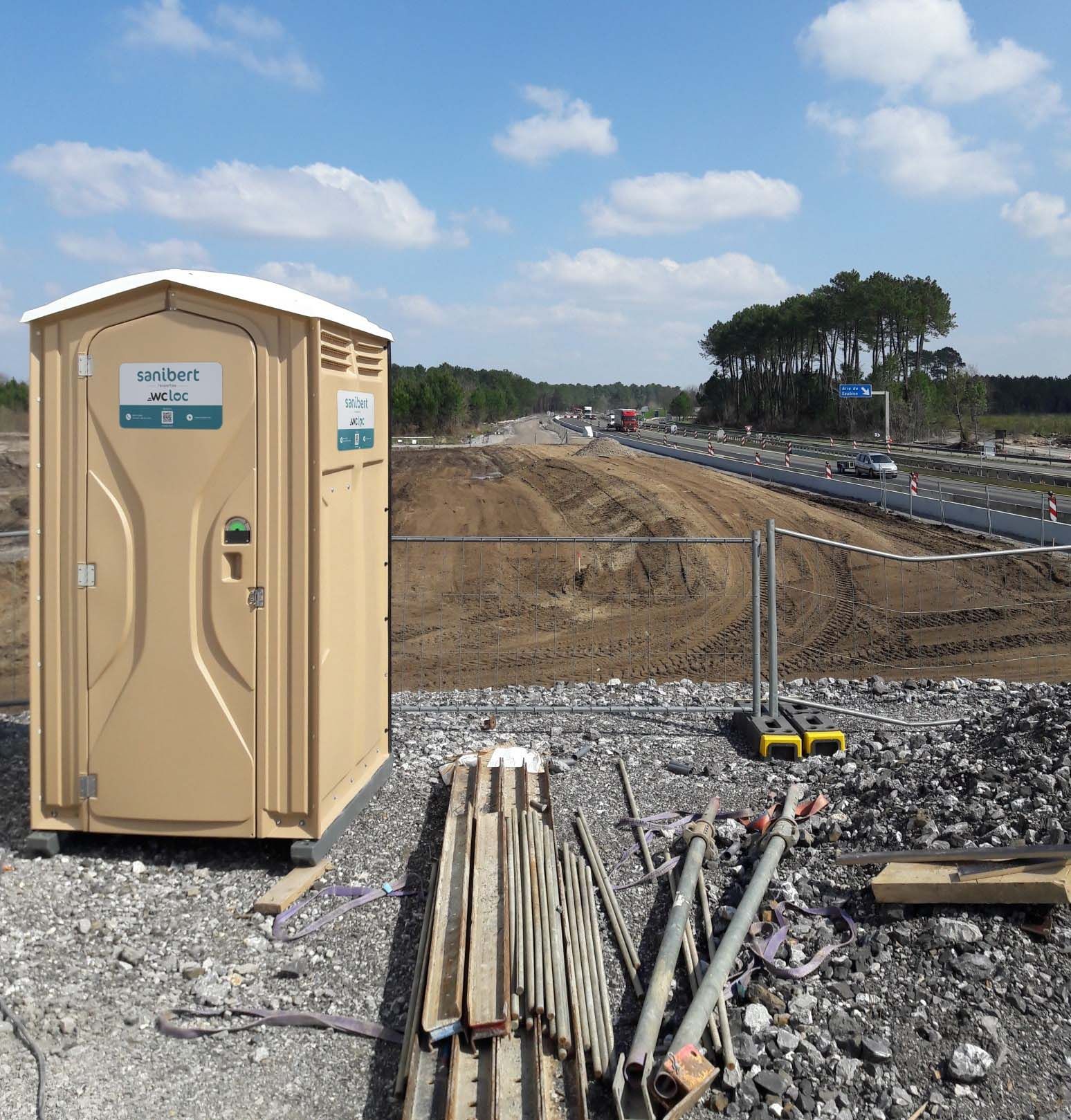 Toilettes portatives beiges sur une surface de gravier avec des matériaux de construction devant une autoroute en construction par une journée ensoleillée.