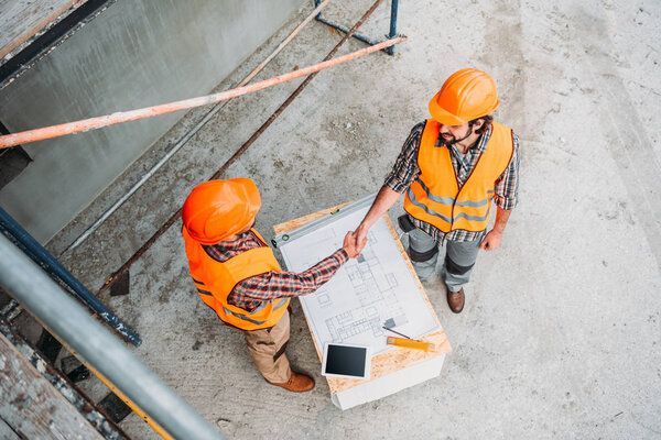Deux ouvriers du bâtiment portant des gilets et des casques orange se serrent la main au-dessus d'un plan sur une surface en béton.