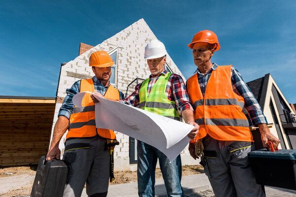 Des ouvriers du bâtiment examinent des plans sur un chantier ; gilets de sécurité et casques aux couleurs vives.