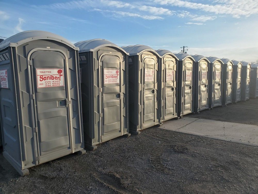 Une rangée de toilettes portables grises sur du gravier sous un ciel bleu avec quelques nuages.
