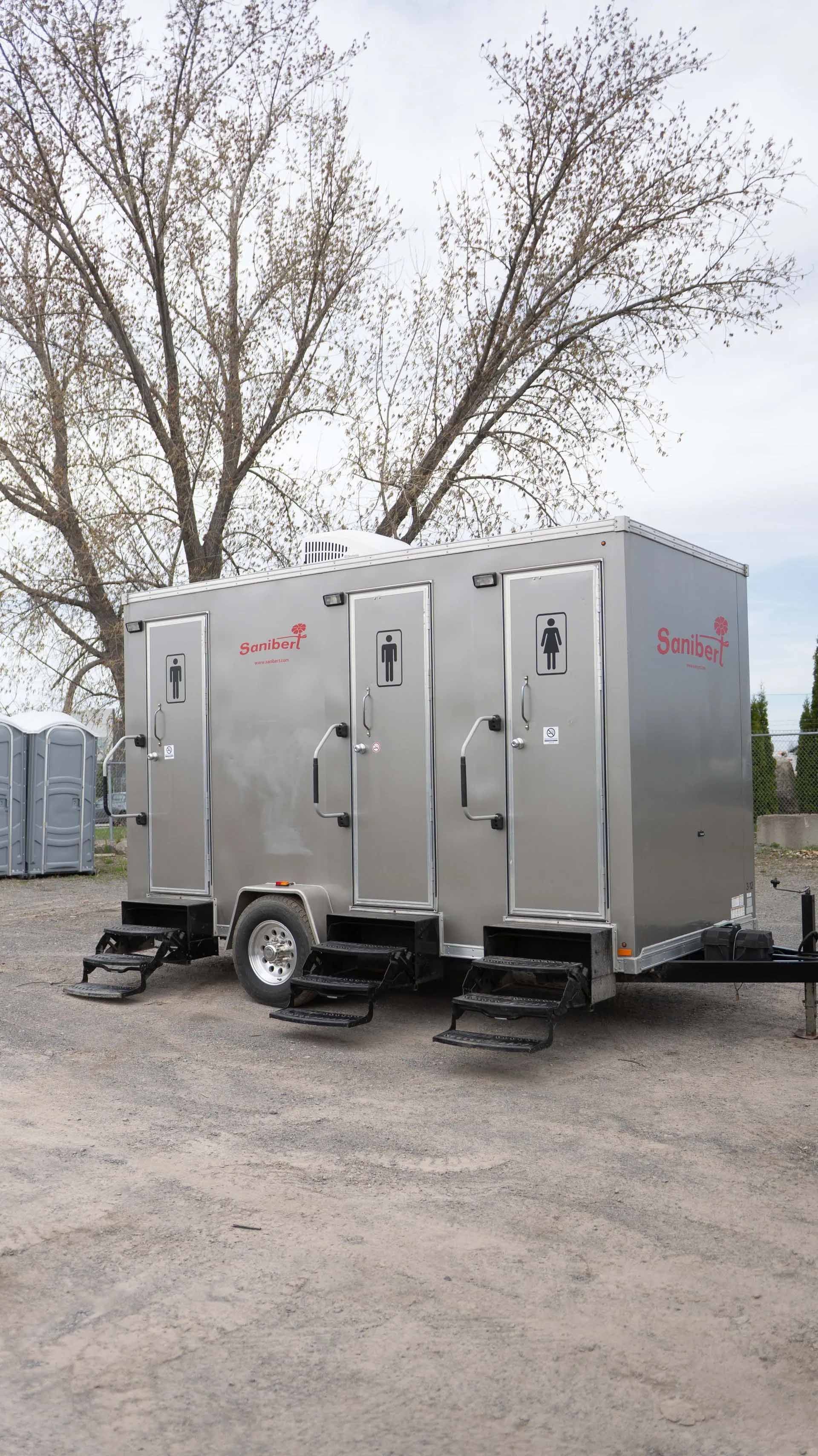 Toilettes mobiles sur un terrain en gravier. Avec plusieurs cabines, un revêtement argenté et des marches. Arbres en arrière-plan.