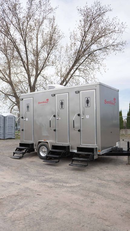 Toilettes mobiles sur un terrain en gravier. Avec plusieurs cabines, un revêtement argenté et des marches. Arbres en arrière-plan.