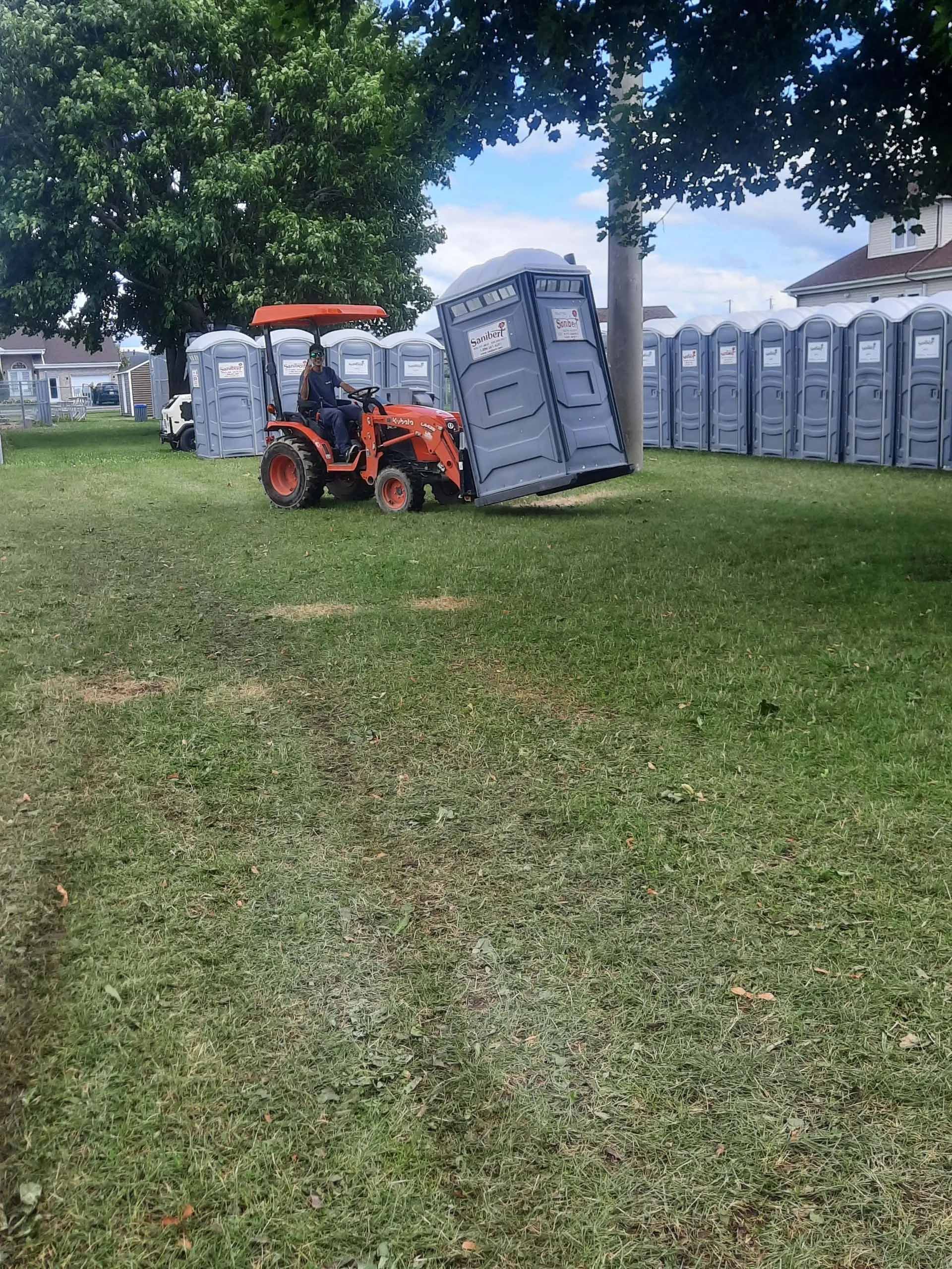 Un tracteur déplace des toilettes portables, avec plusieurs autres alignées sur un champ herbeux, sous un arbre.