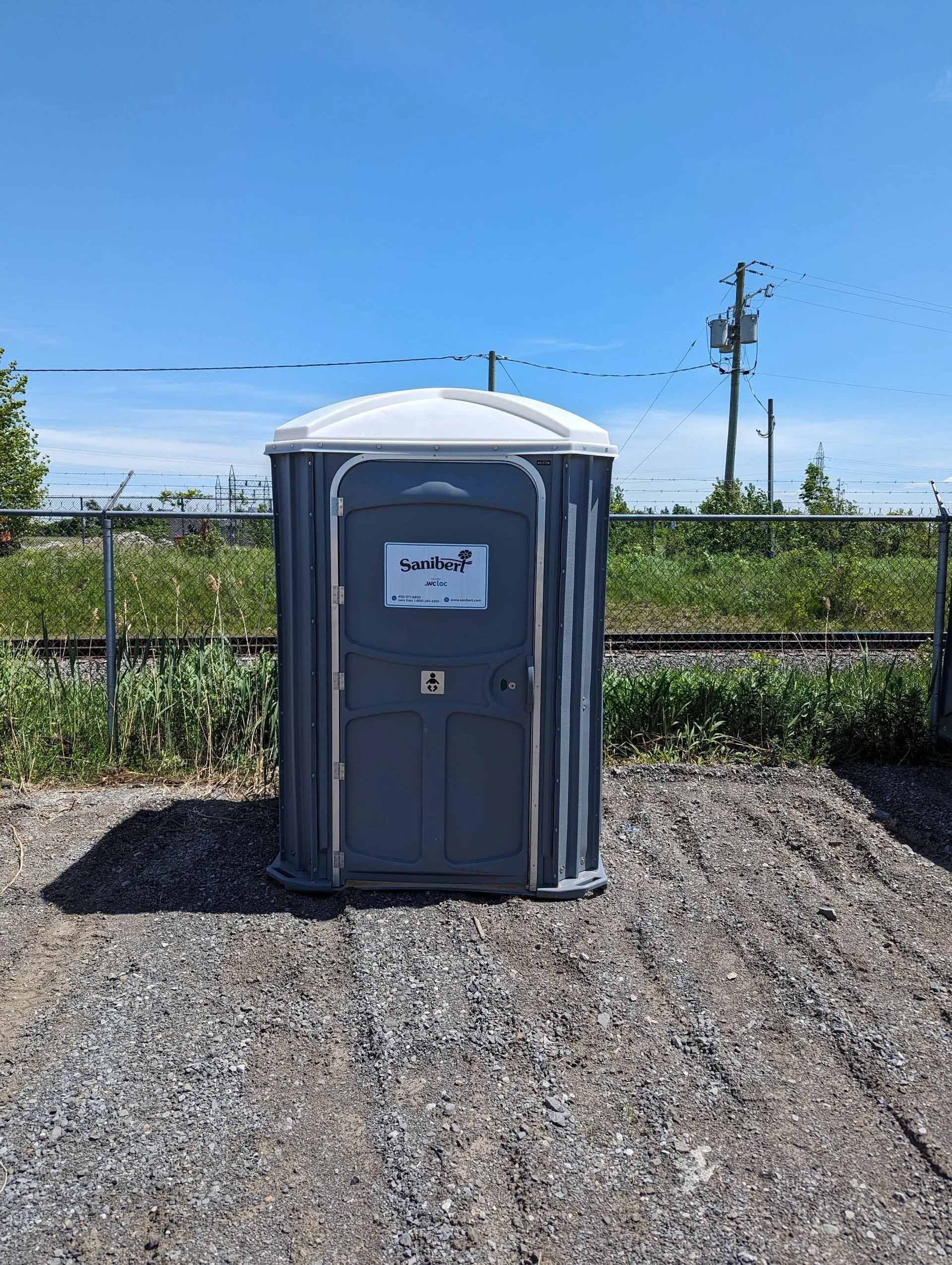 Toilettes portables grises sur gravier à côté d'une voie ferrée sous un ciel bleu.