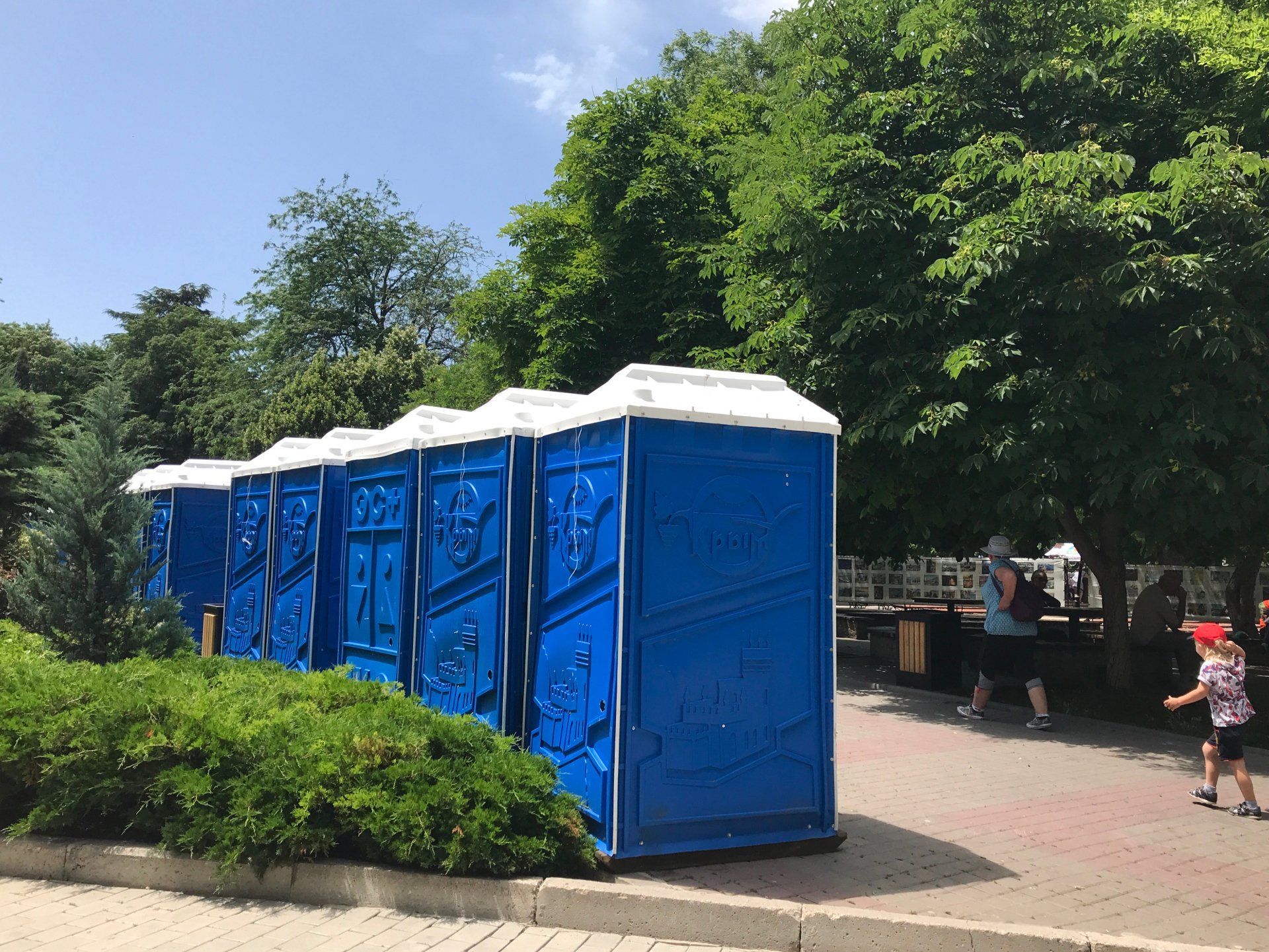 Toilettes portables bleues en rangée, situées près de la verdure avec des personnes marchant dans un parc.