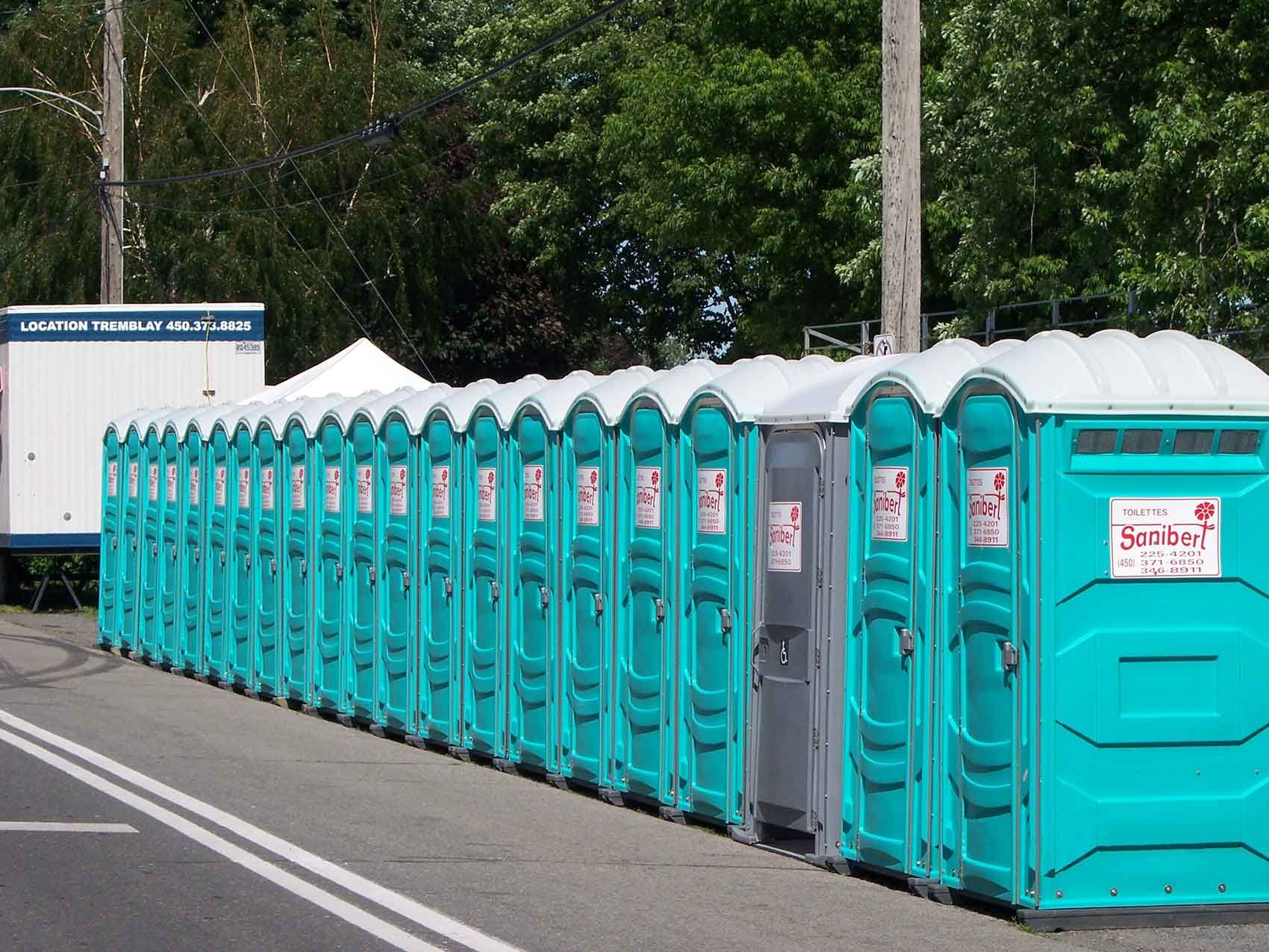 Rangée de toilettes portables turquoise alignées sur une surface pavée, avec une remorque blanche en arrière-plan.