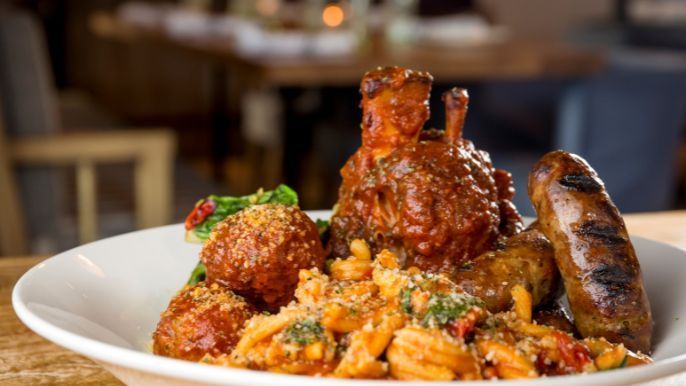 A plate of pasta with meatballs, a grilled sausage, and braised meat, set in a restaurant.