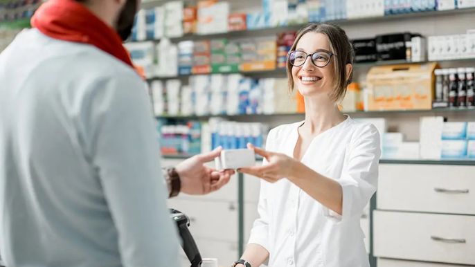 Pharmacist handing medication box to a customer in a pharmacy; the pharmacist smiles.