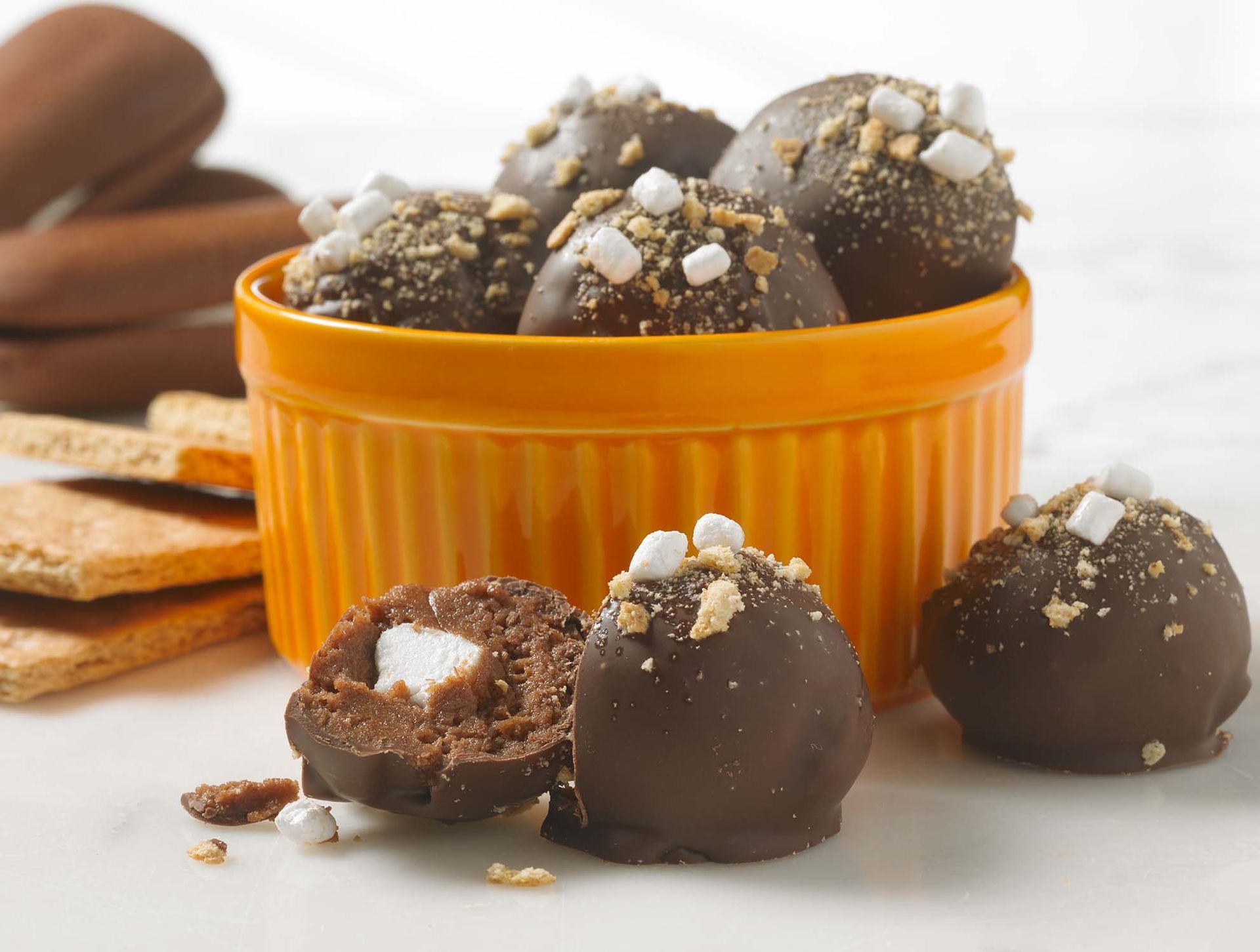 S’mores truffles topped with tiny marshmallows and graham cracker crumbs in an orange ramekin, with crackers on the side.
