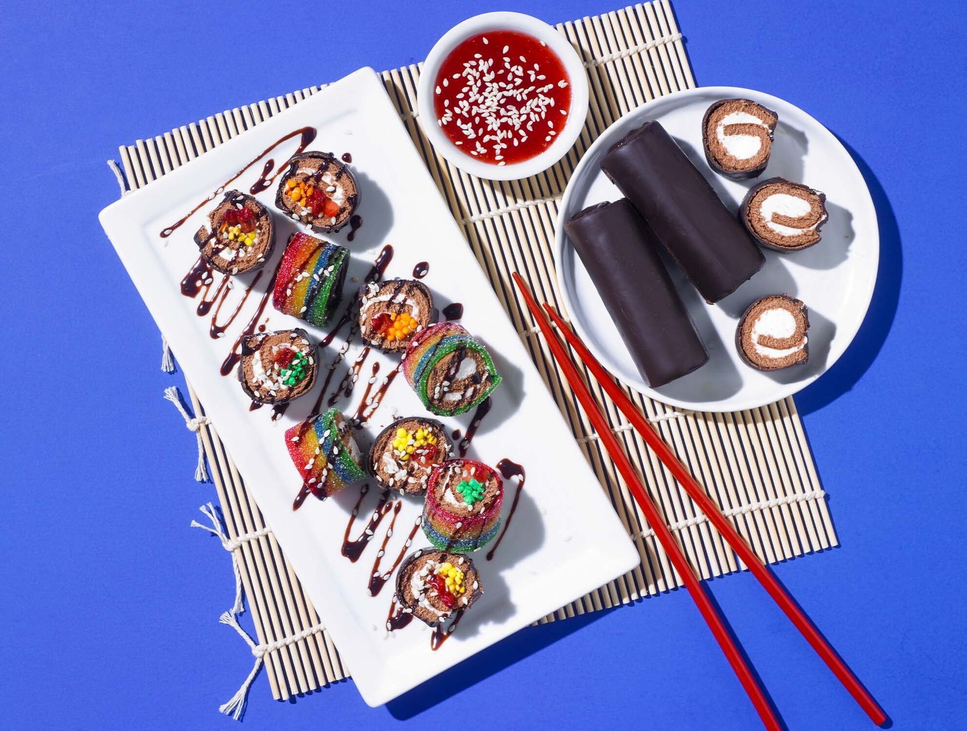Sushi and chocolate dessert rolls on plates with red chopsticks against a bright blue background.