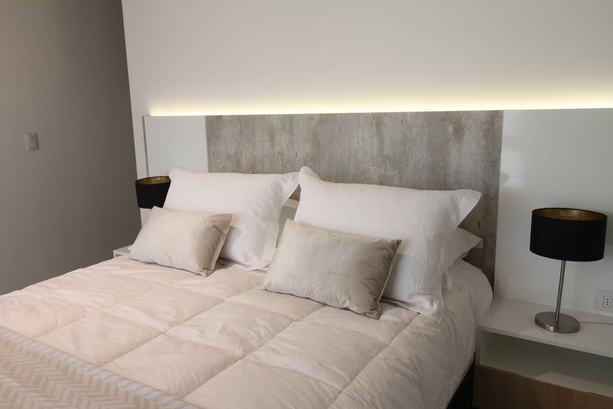 Dormitorio con cama, colores neutros, dos mesitas de noche e iluminación incorporada en el cabecero.