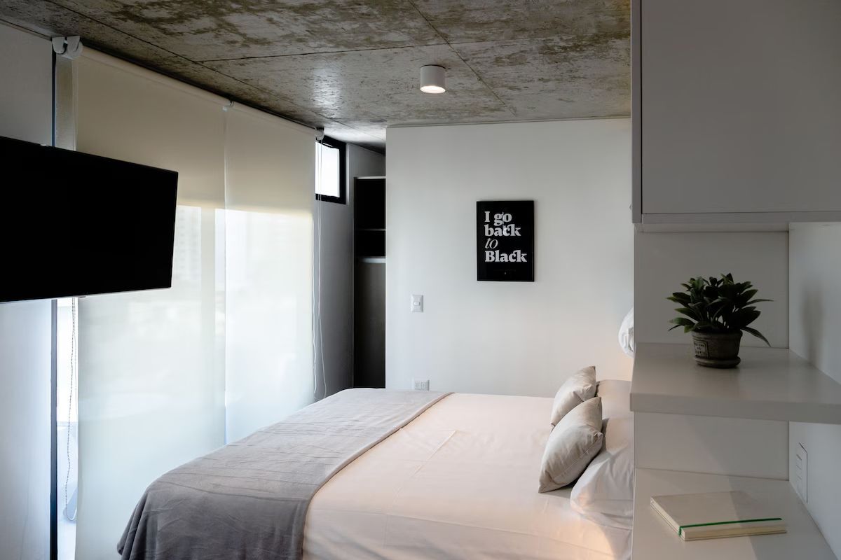 Dormitorio con cama blanca, manta gris, TV y una pequeña planta en un estante.