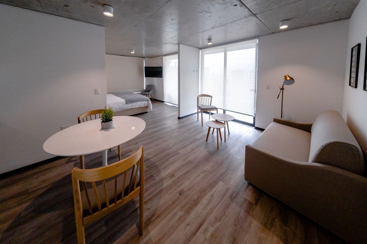 Moderno apartamento tipo estudio con sofá, mesa, sillas, cama y grandes ventanales.