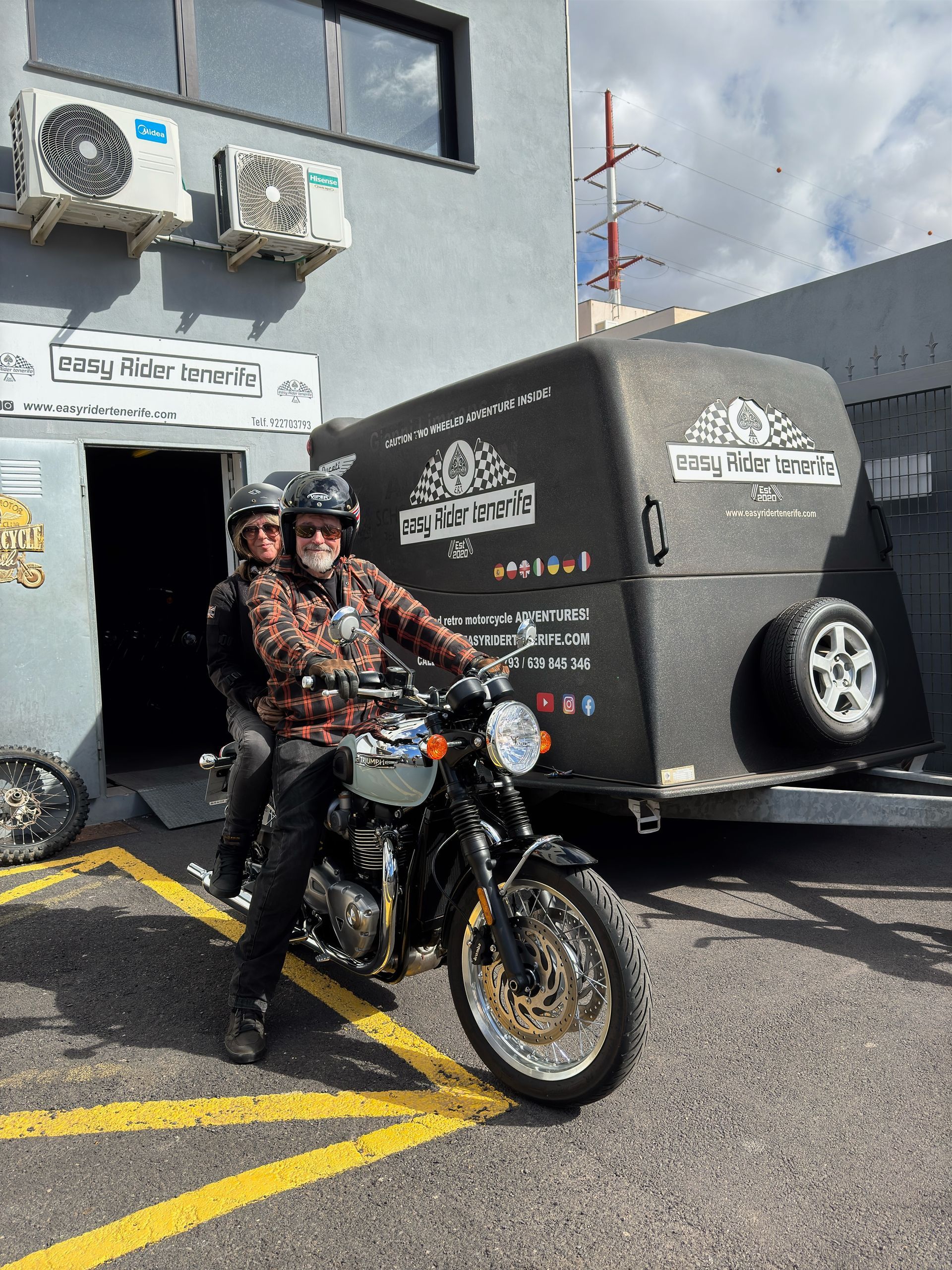 A man is riding a motorcycle next to a trailer in a parking lot.