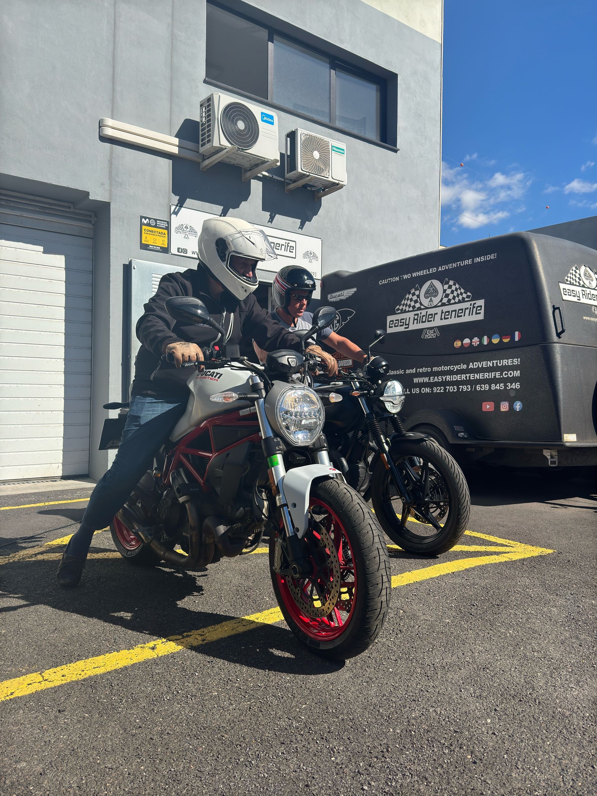 Two men are riding motorcycles in a parking lot in front of a building.