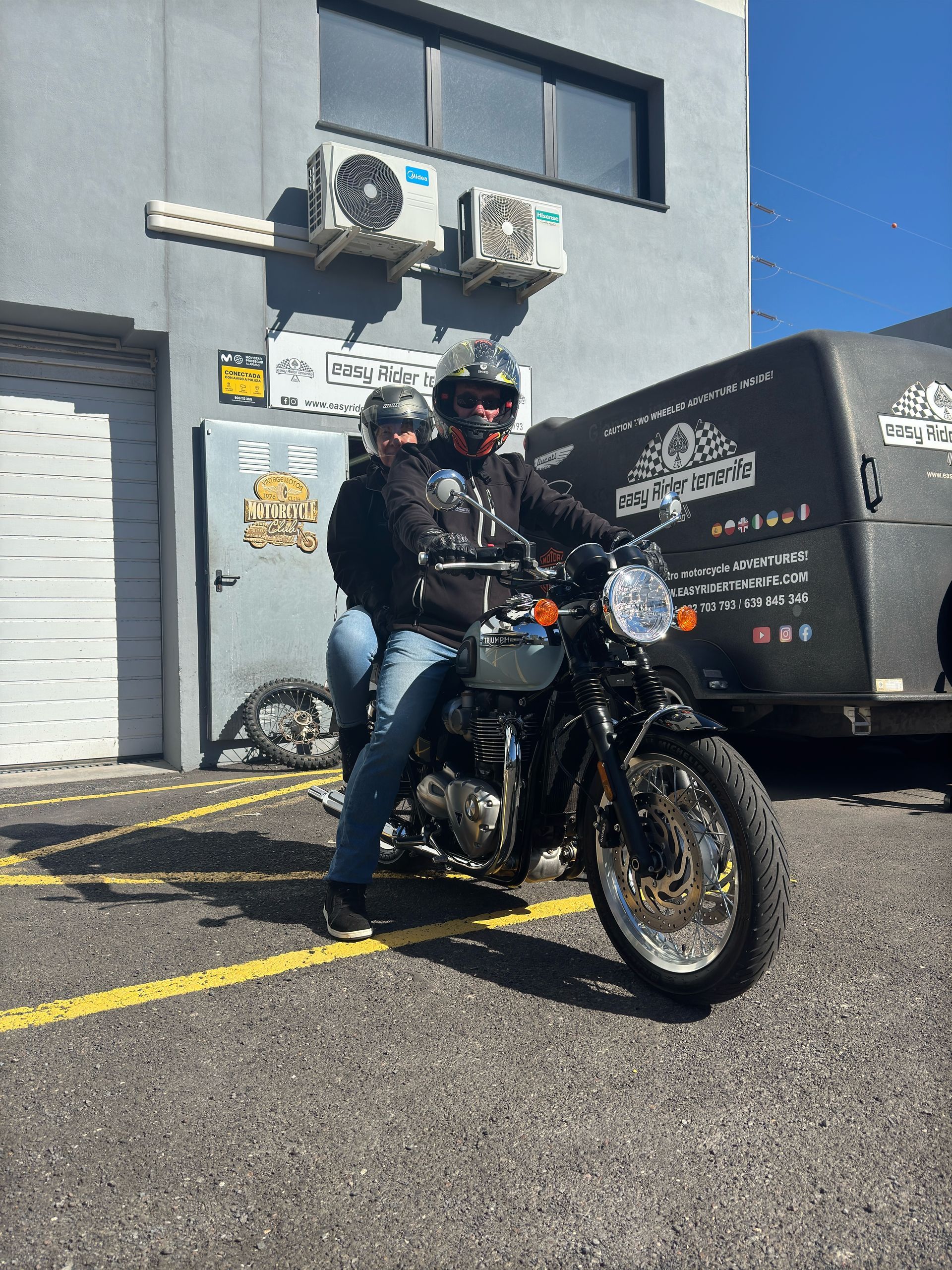 Two men are riding a motorcycle in a parking lot in front of a building.