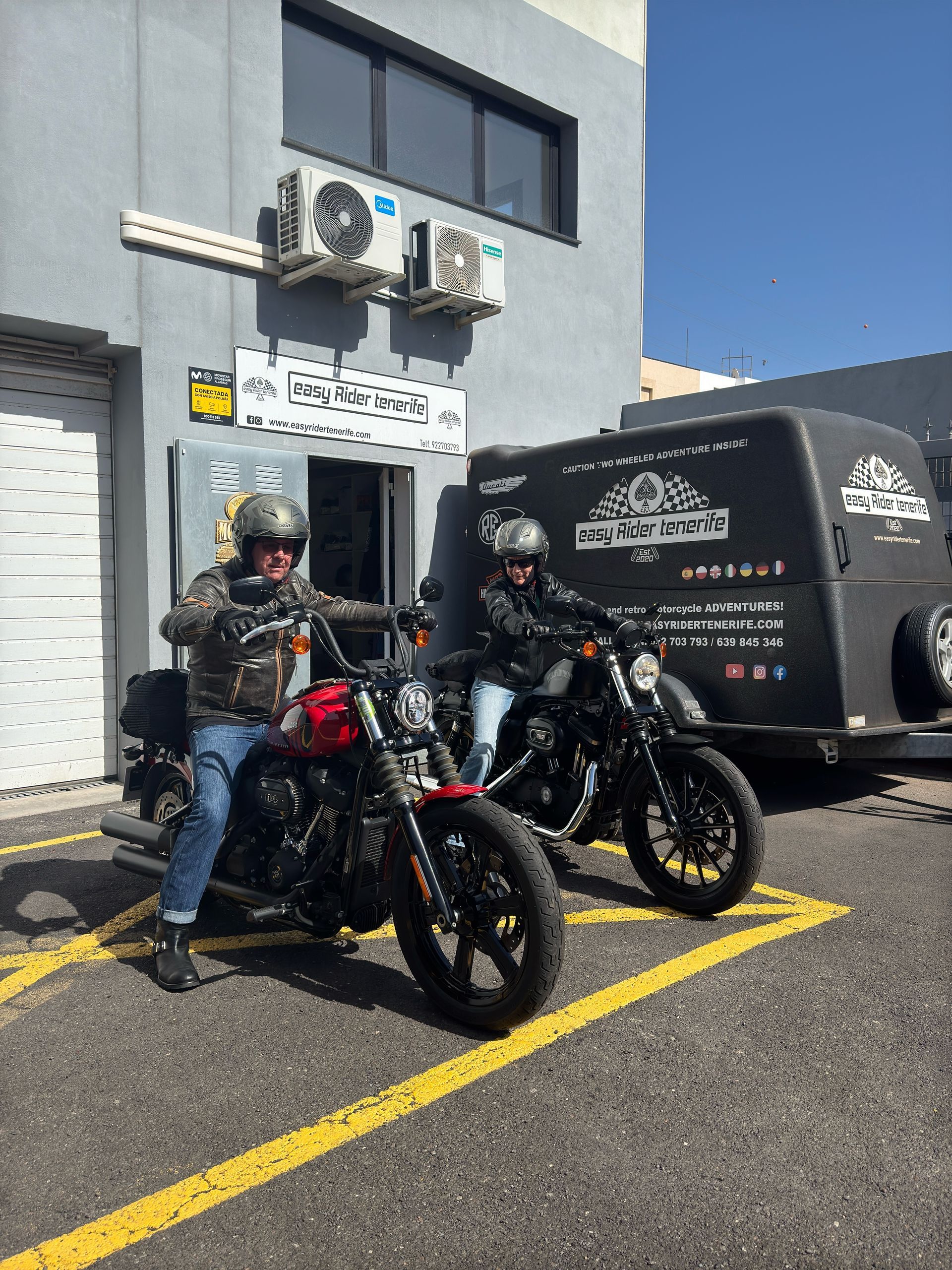 Two men are riding motorcycles in a parking lot in front of a building.