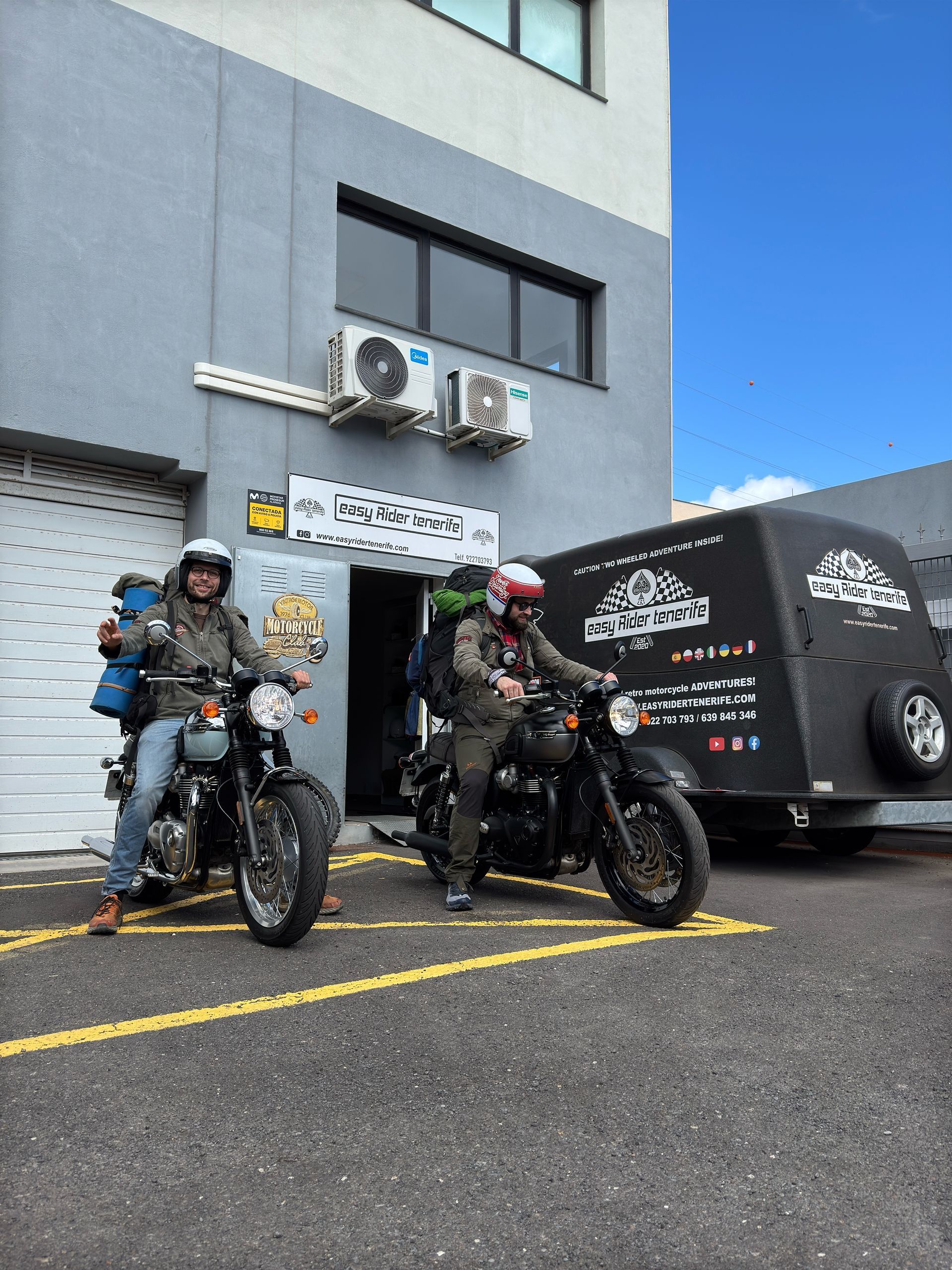 Two men are standing next to their motorcycles in front of a building.