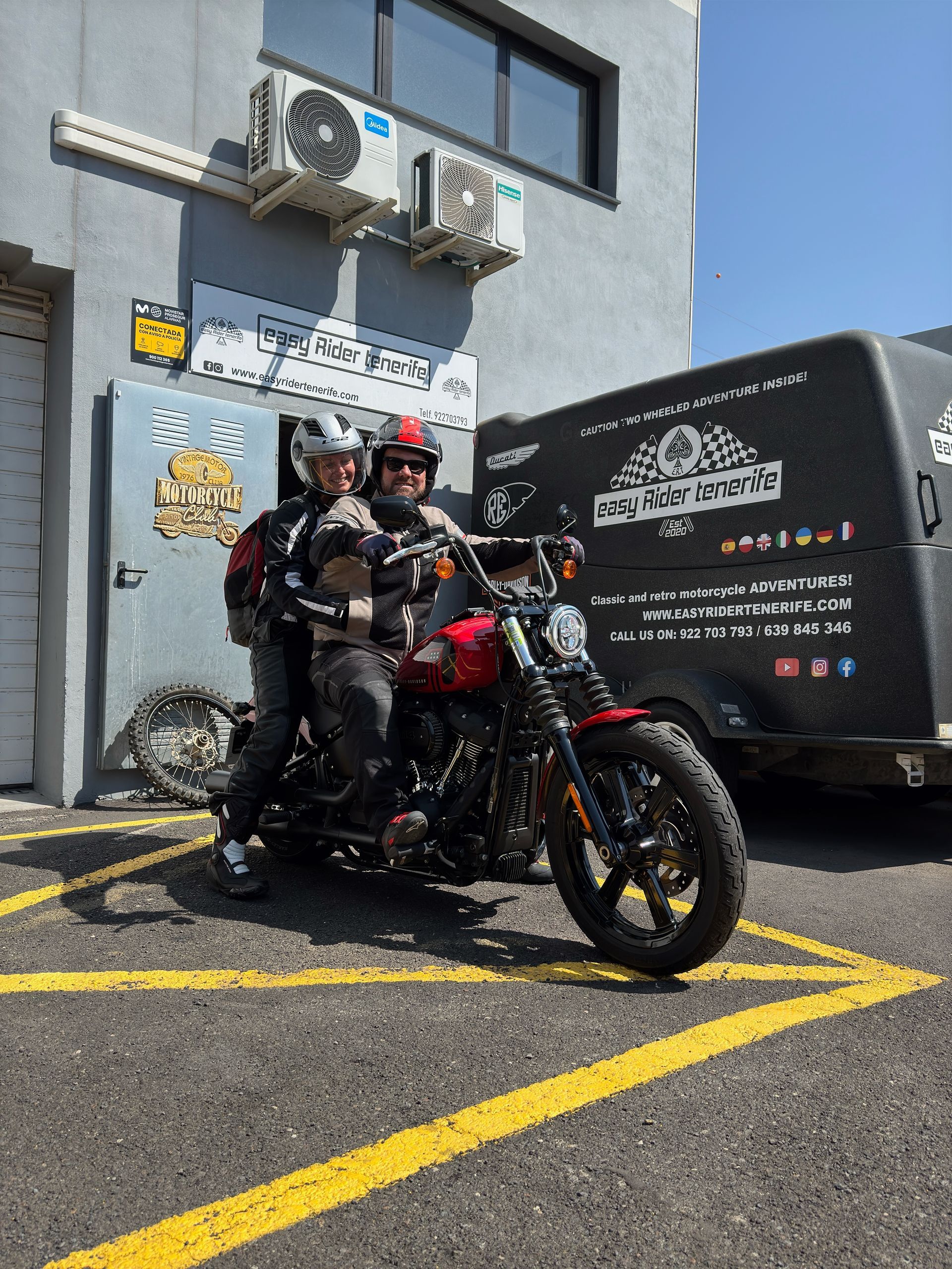 Two people are riding a motorcycle in a parking lot in front of a building.