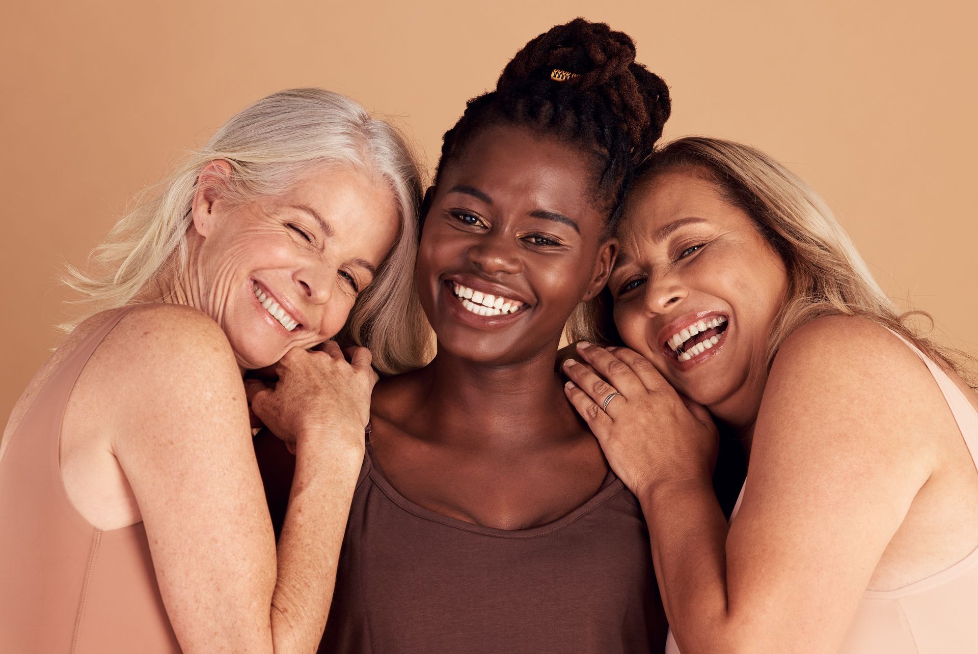 Three smiling people of diverse skin tones embrace, posing in front of a neutral backdrop.